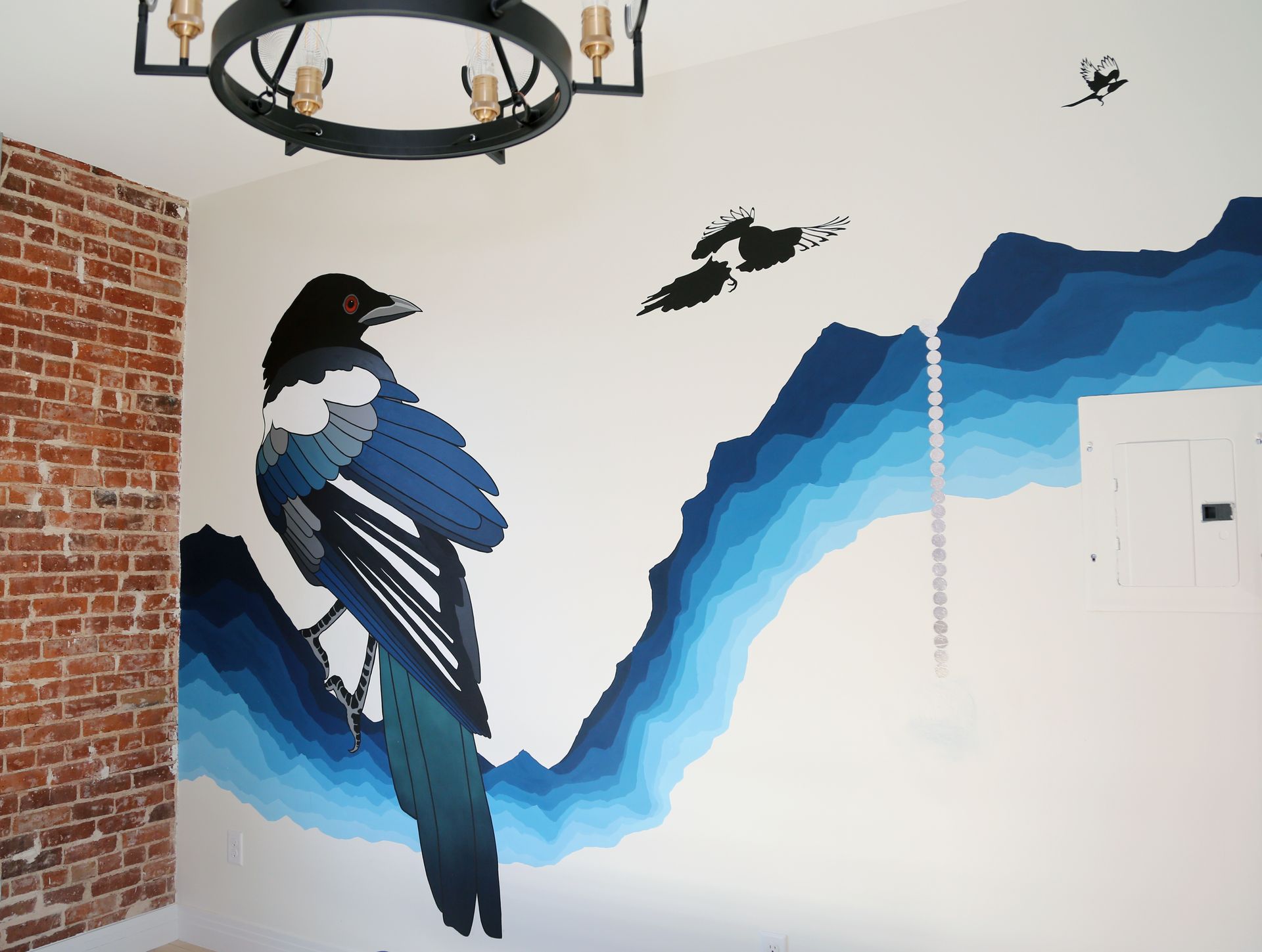 Mural of a blue-and-black bird on a white wall beside exposed brick and a hanging chandelier.