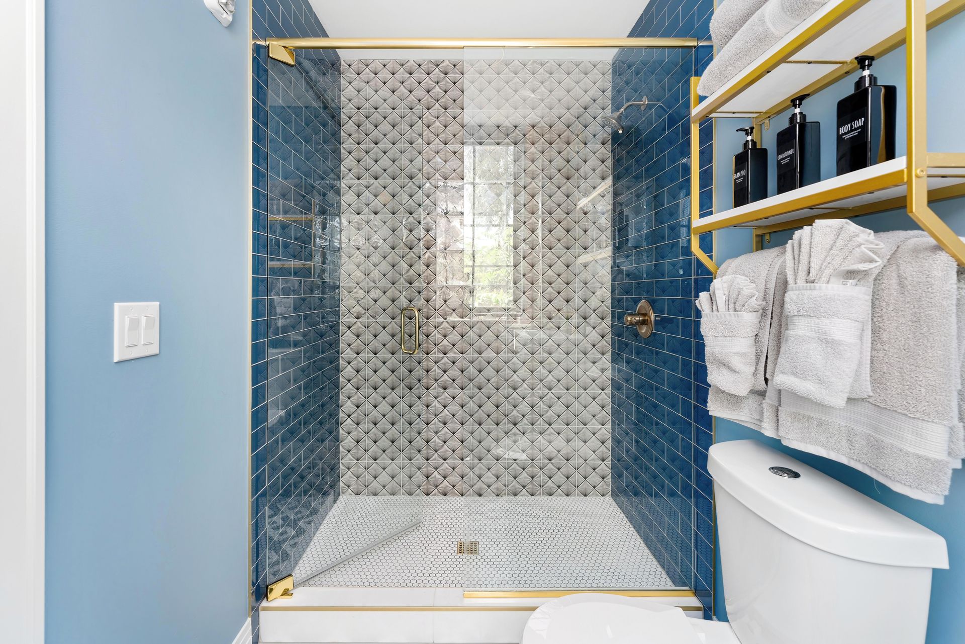 Blue-tiled bathroom with a glass shower, white towels, and a toilet beside yellow shelving