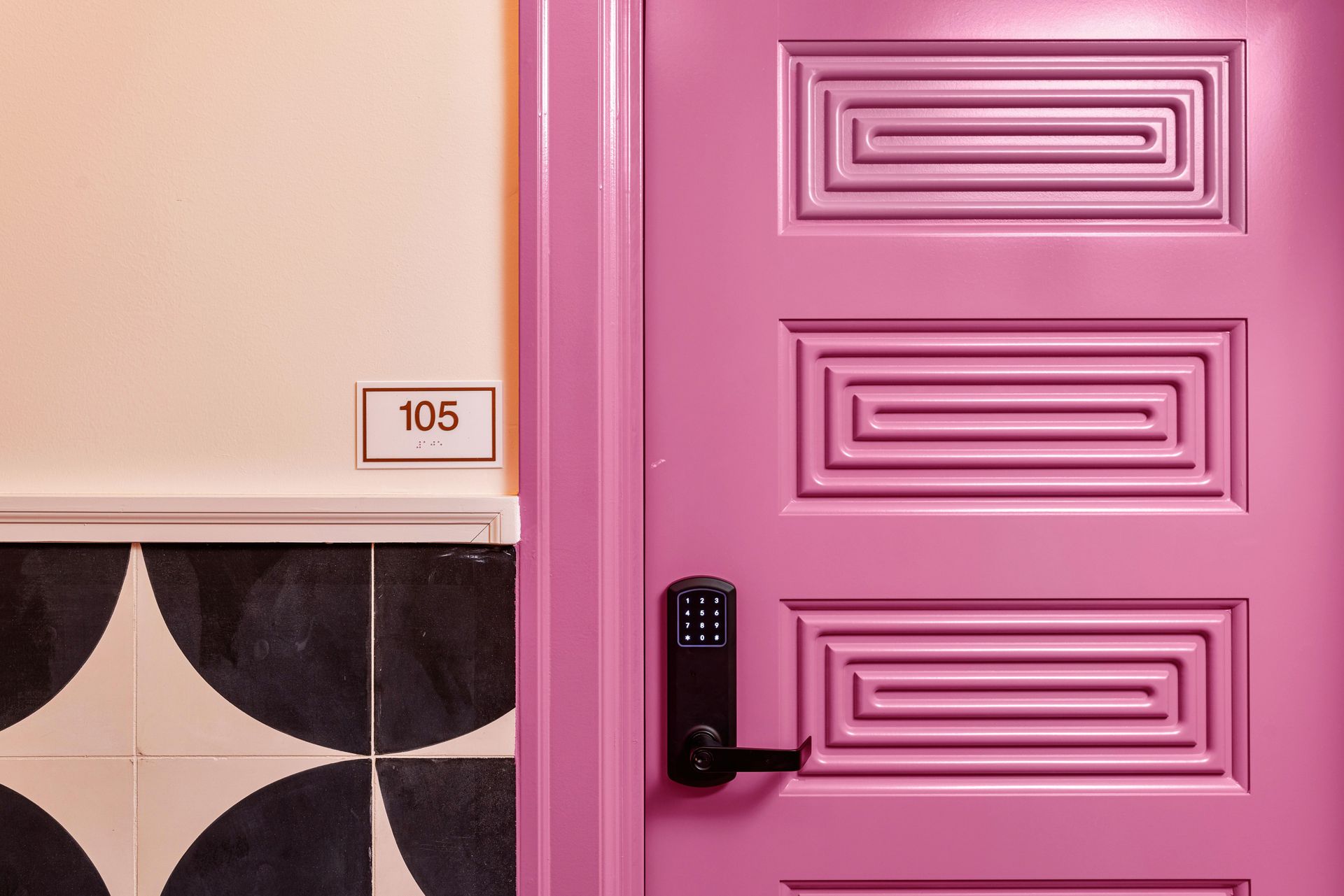 Pink door with black keypad lock beside a room number 105 sign and patterned wall tiles