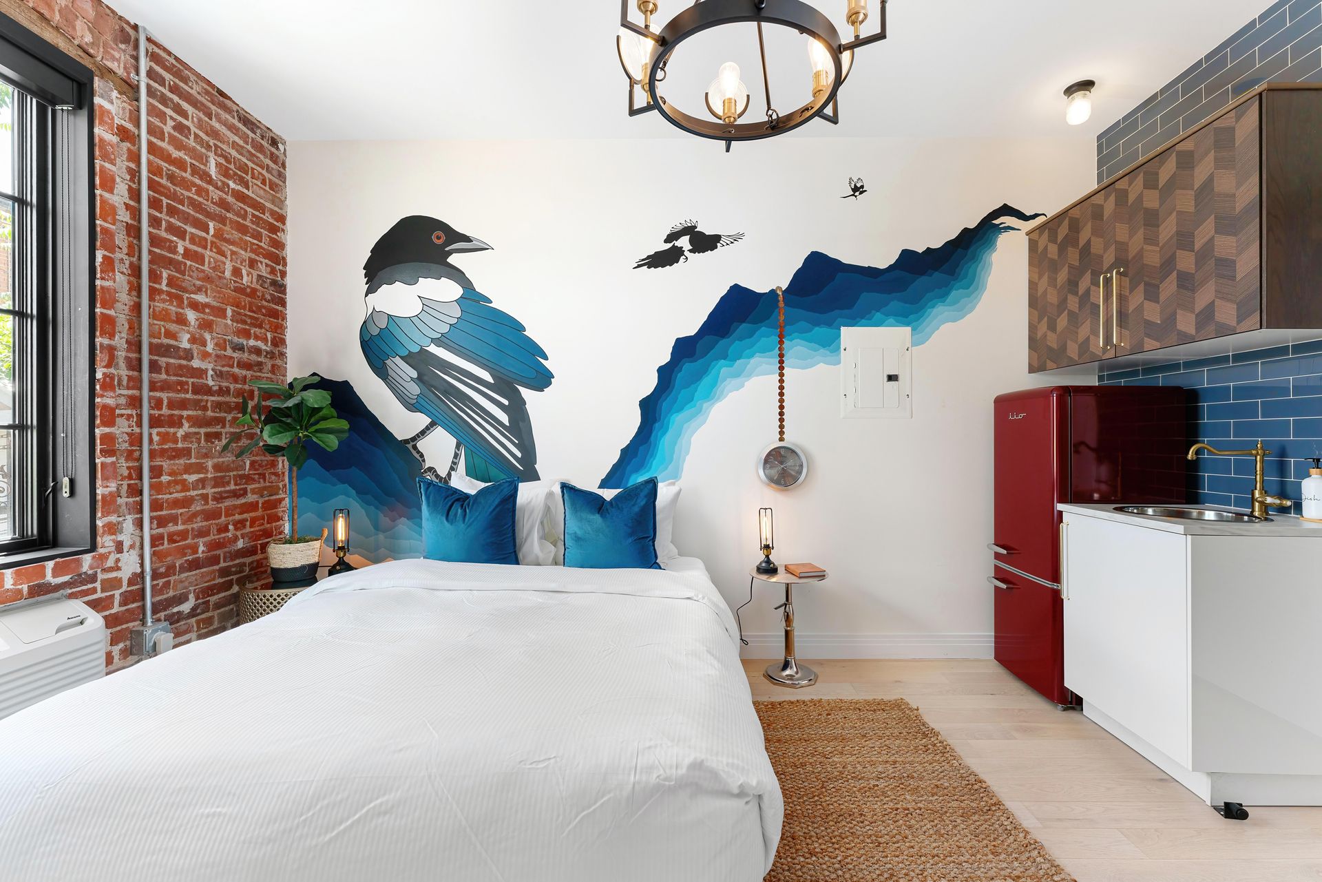 Bedroom with mural of a blue bird over bed, brick wall, red fridge, and small kitchenette