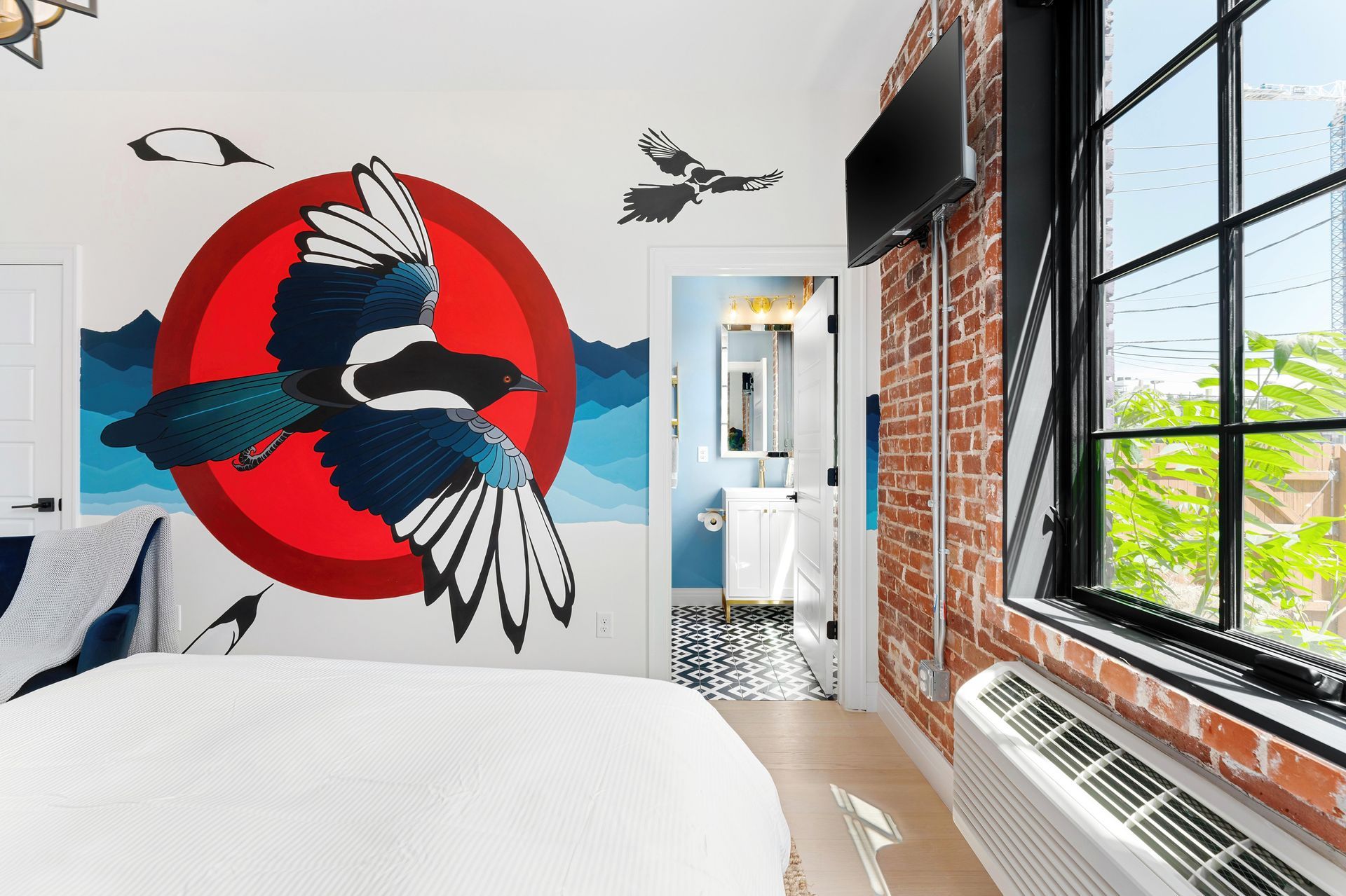 Bright bedroom with bird mural, red sun, and large brick-walled windows overlooking greenery.