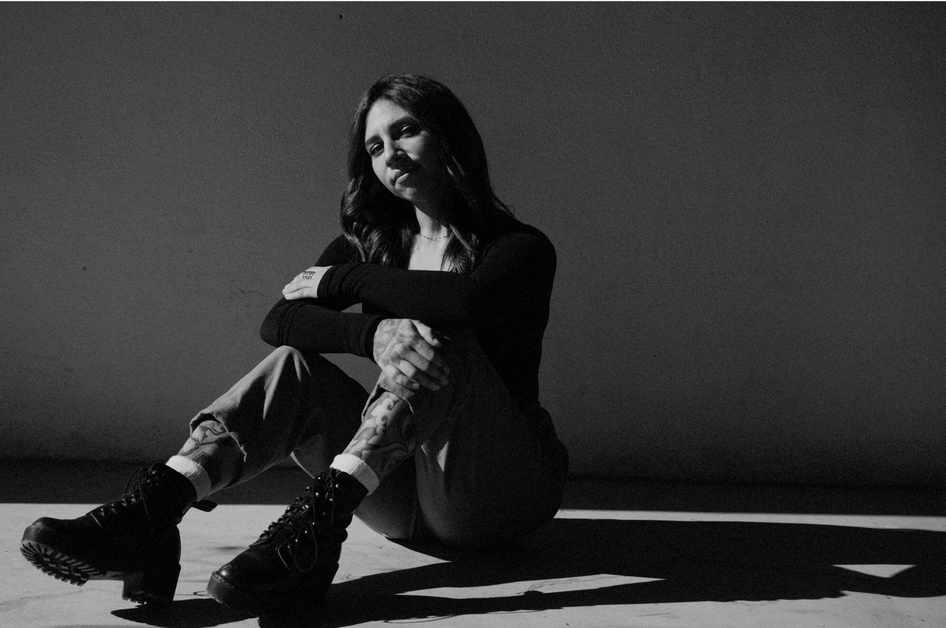 Black-and-white photo of a seated person in boots, hugging their knees in a stark, shadowed room