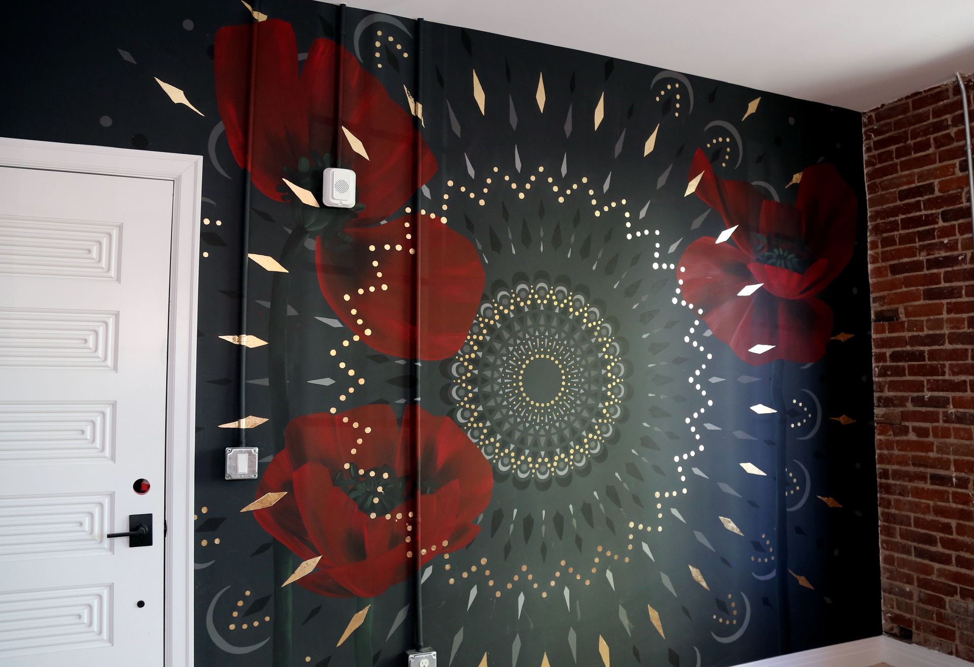 Mural with red flowers and a green circular pattern on a dark wall beside a white door and brick accent wall