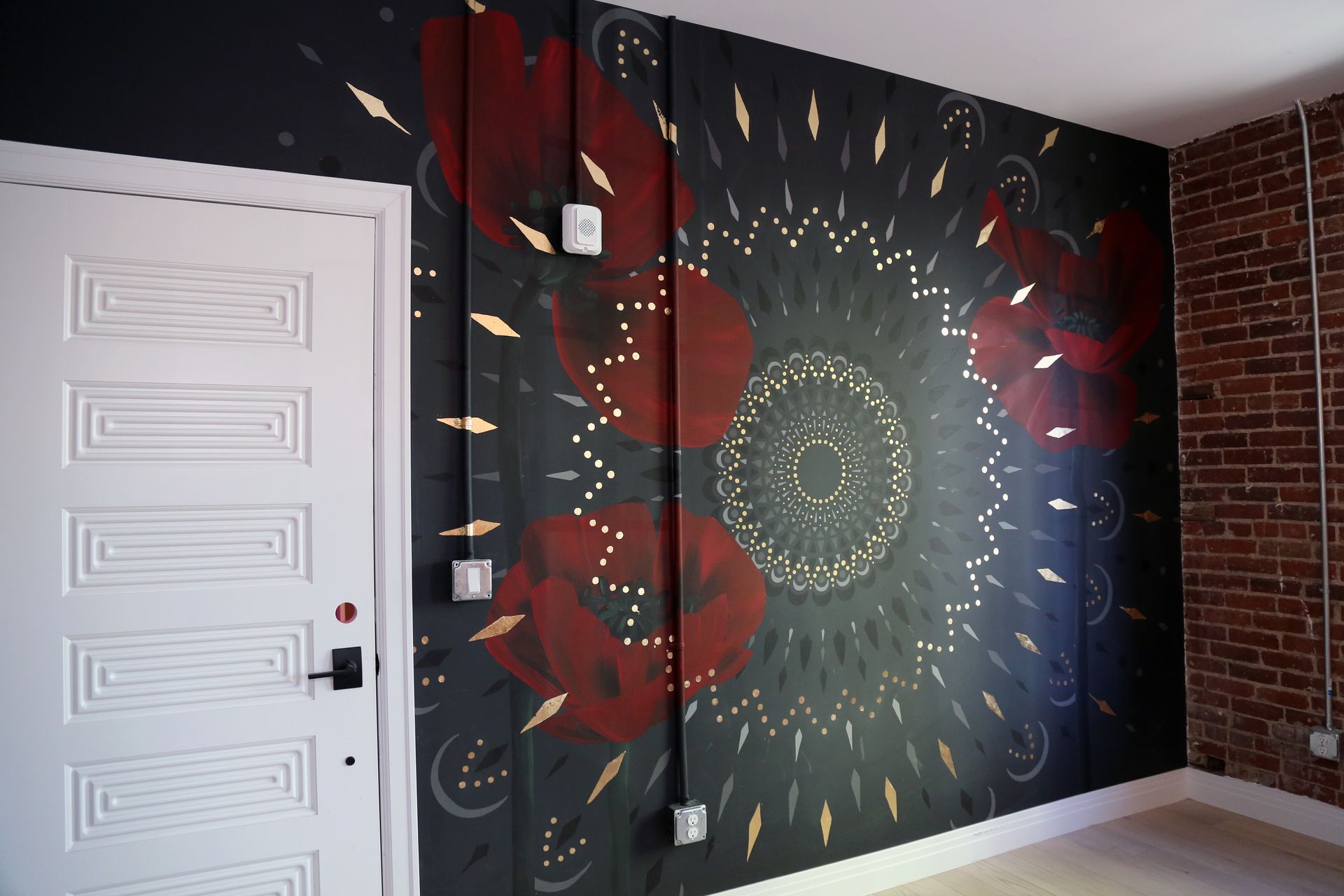 Dark mural with red circles and a central sunburst on a black wall beside a white door and exposed brick.