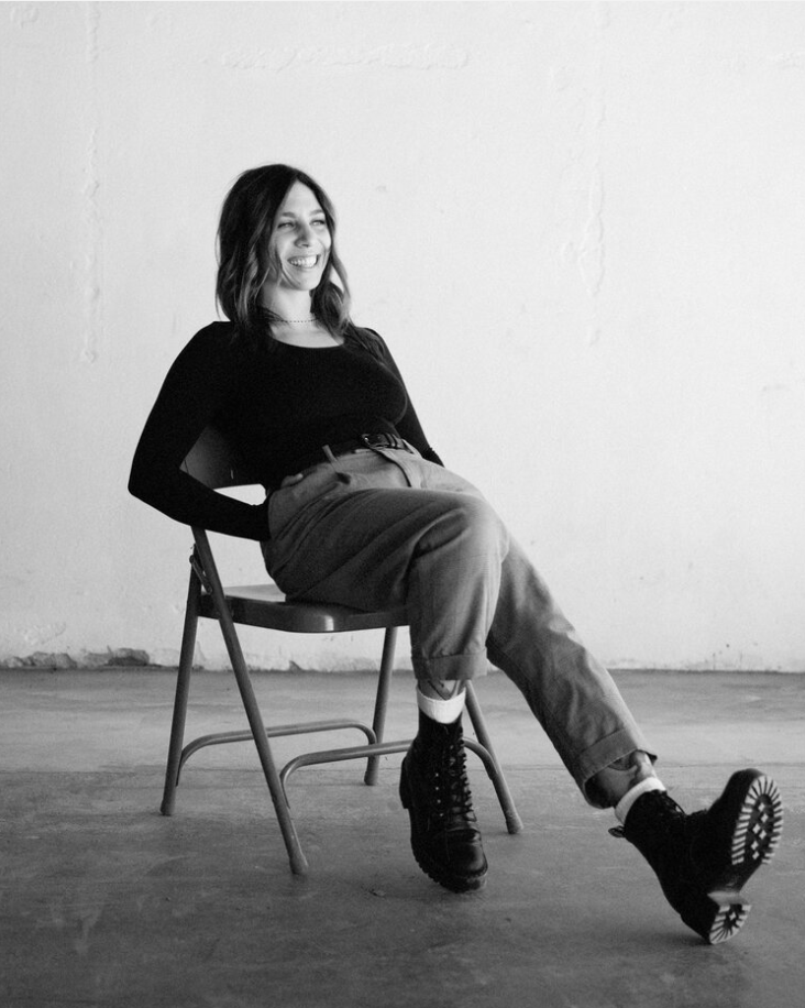 Woman reclining on a chair in a black top and pants, smiling in a minimalist studio setting