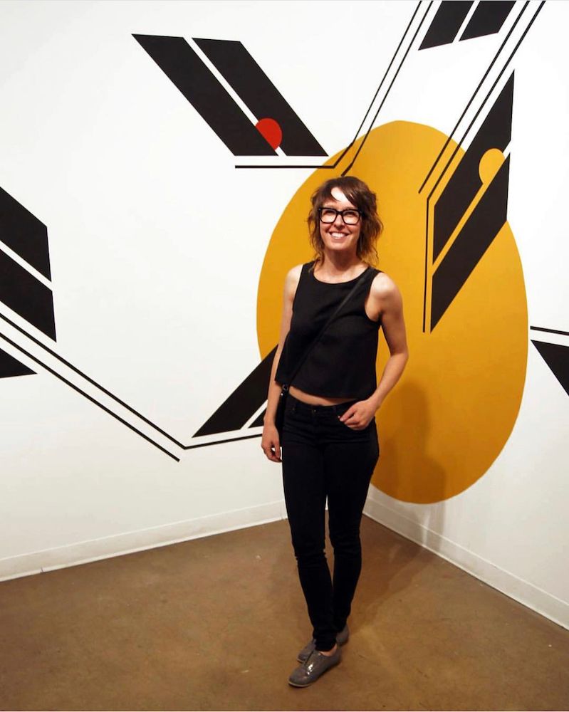 Woman in black posing in front of a bold black, white, and yellow abstract wall mural.