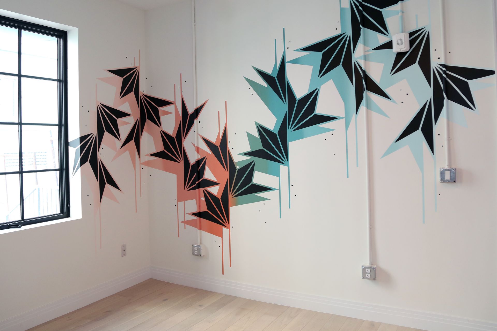 Abstract black, teal, and coral geometric wall mural in a white room with a window and outlets