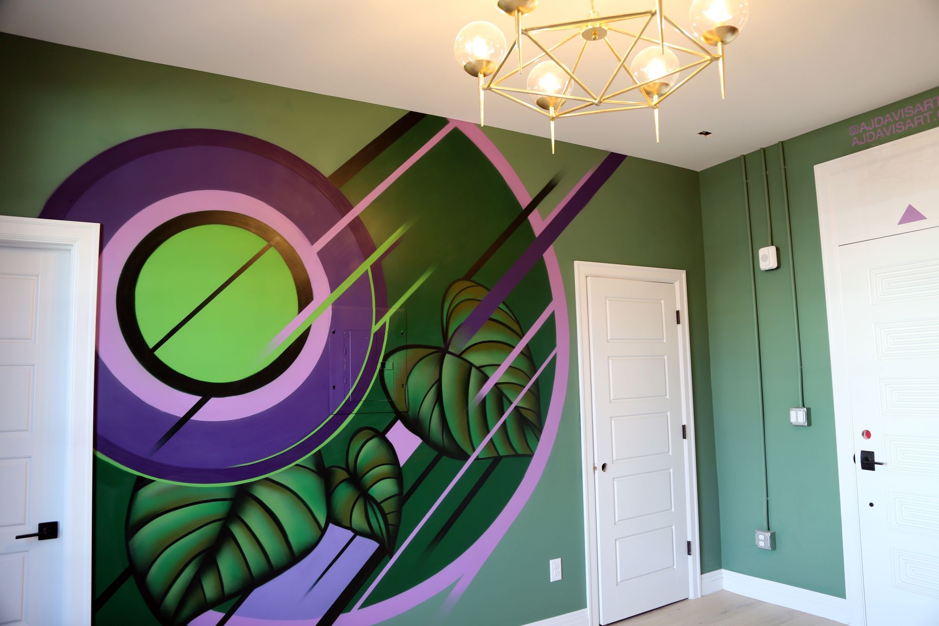 Colorful mural on a bedroom wall with green and purple abstract leaf designs, white doors, and a chandelier.