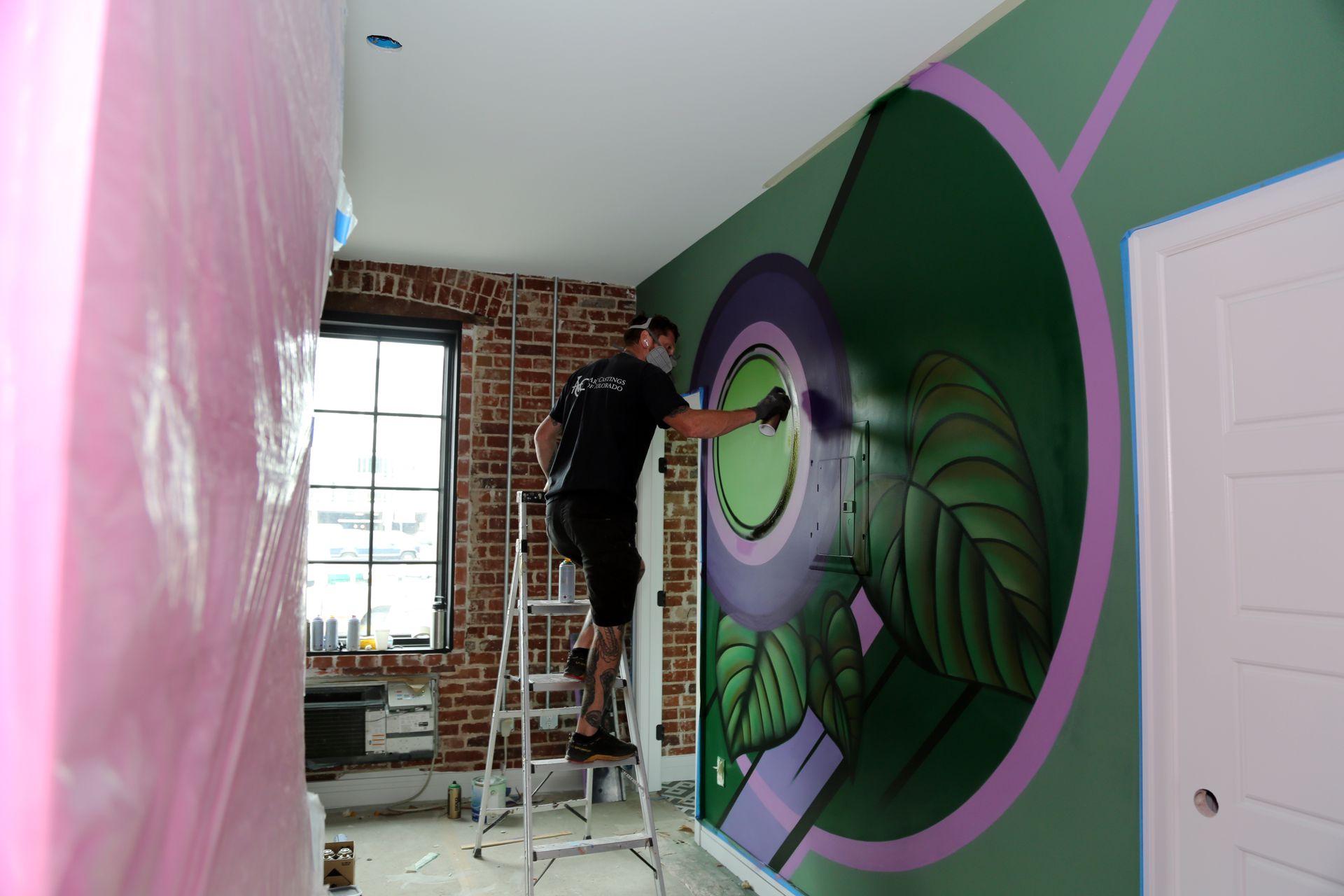 Artist painting a large green mural on a wall in a room with exposed brick and pink-painted walls