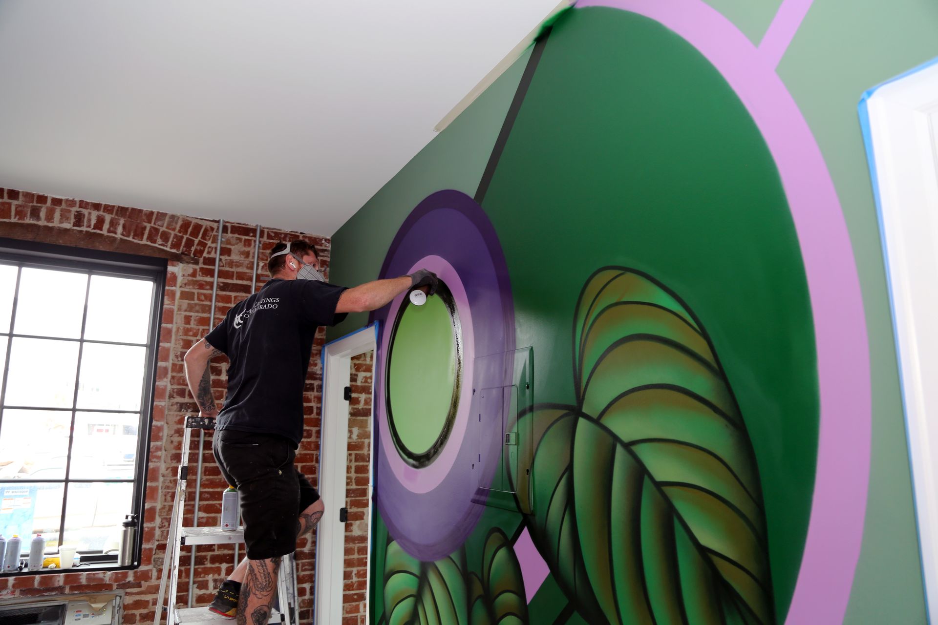Person painting a purple-and-green mural on an indoor wall near a window