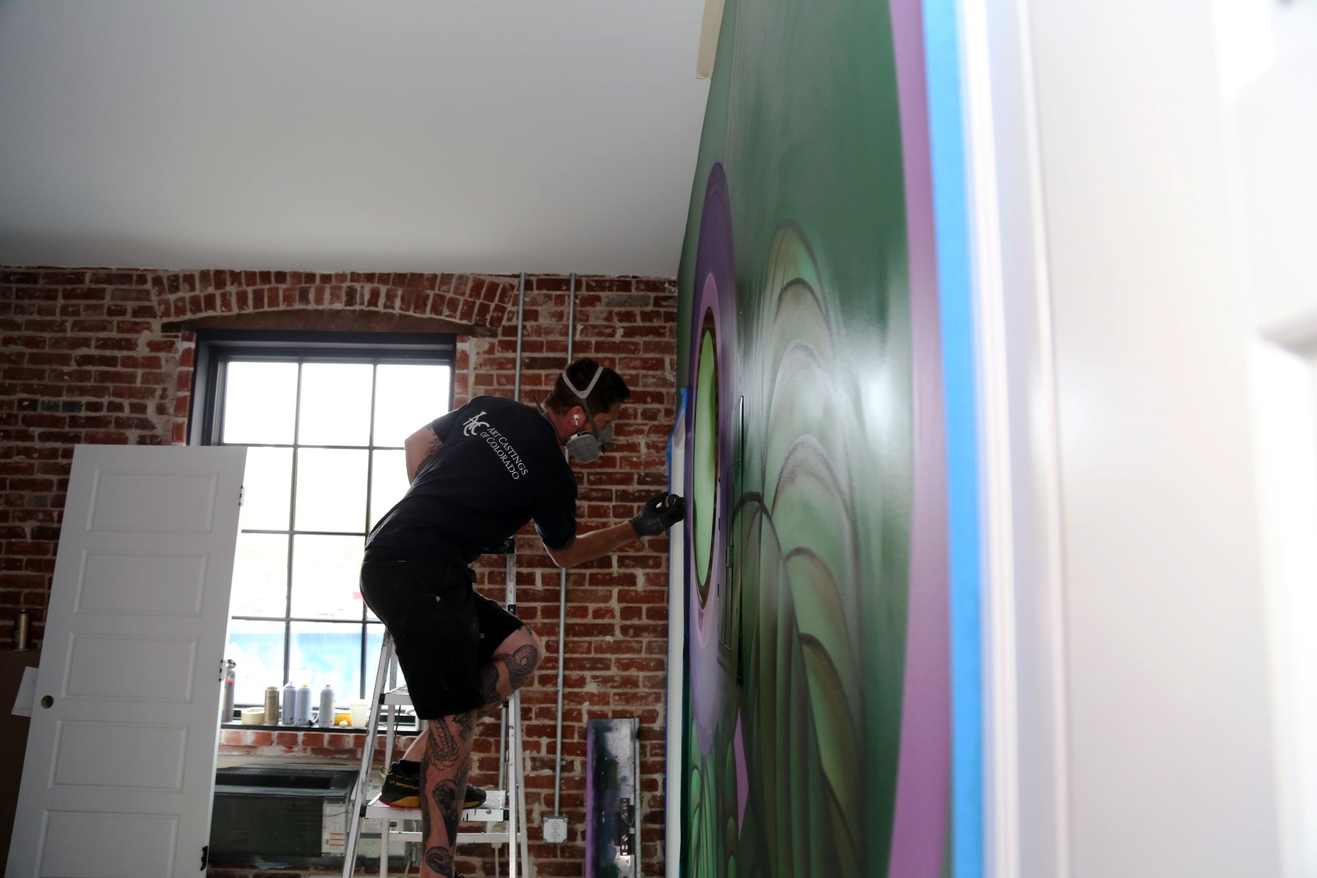 Person painting a mural on a wall inside a room with exposed brick and a ladder