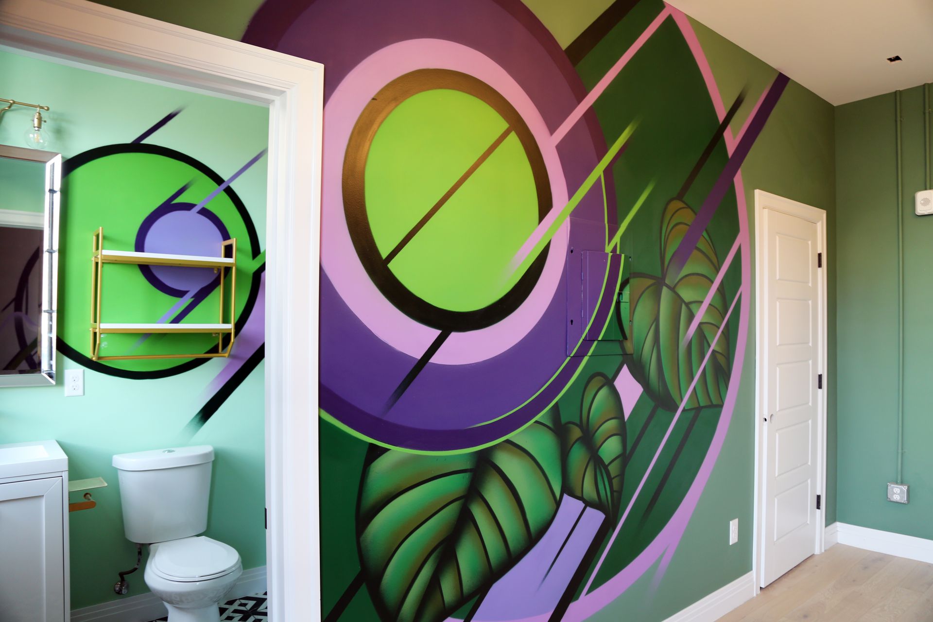 Colorful abstract mural on a wall beside a small bathroom with toilet and sink