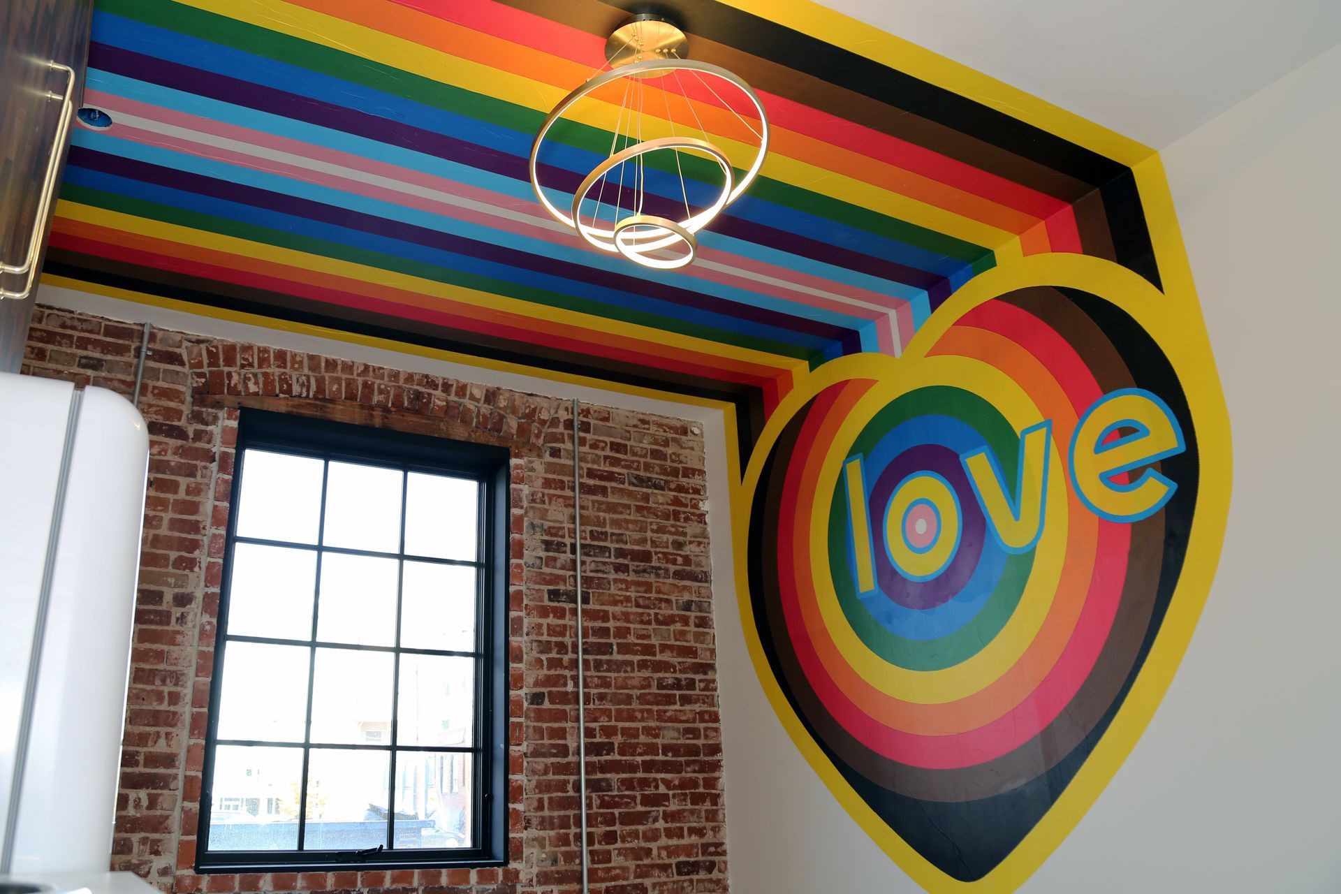 Rainbow wall mural with a heart and “love” text in a room with a window and exposed brick.