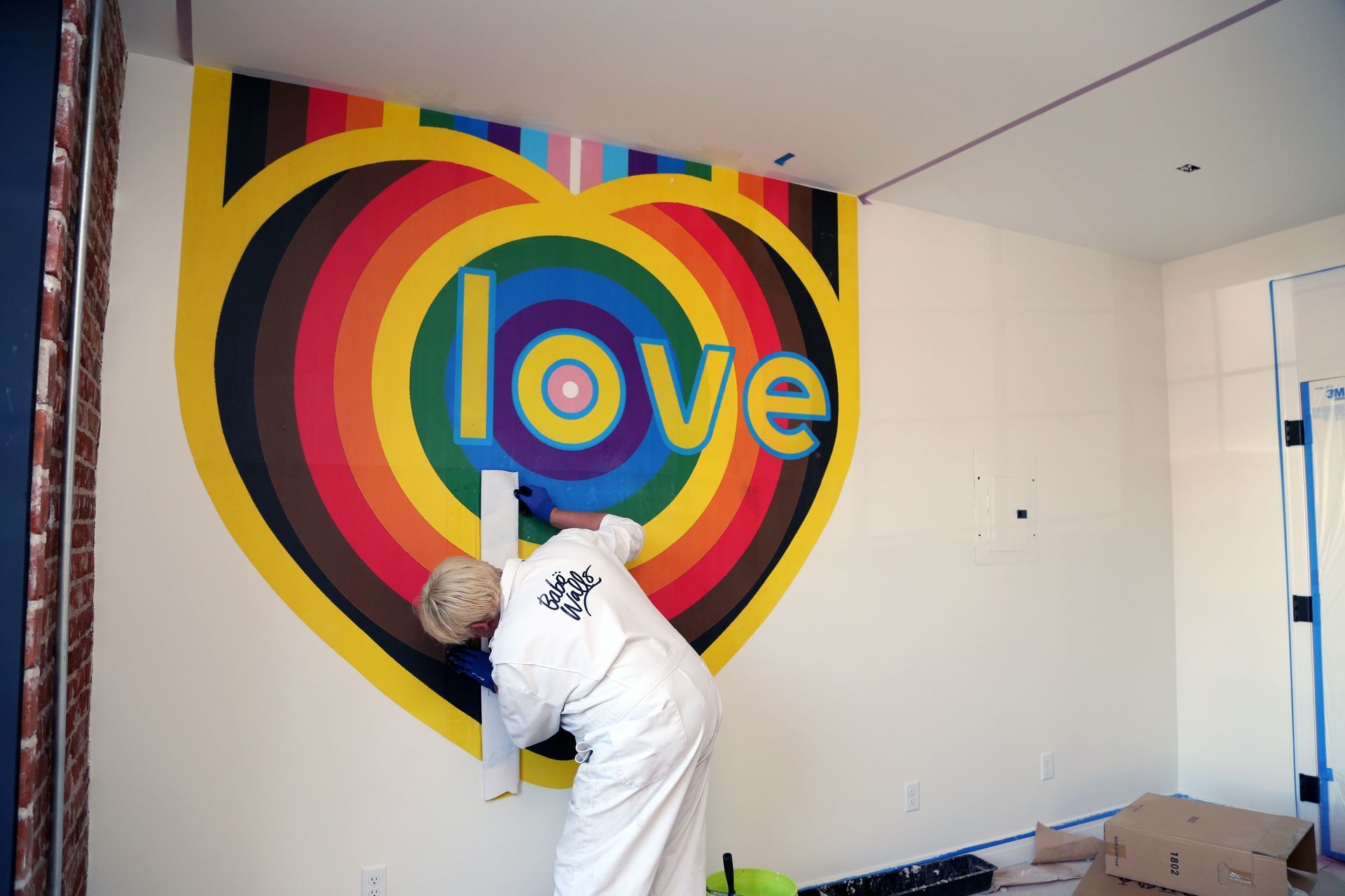 Person painting a colorful heart mural with the word “love” on a wall indoors