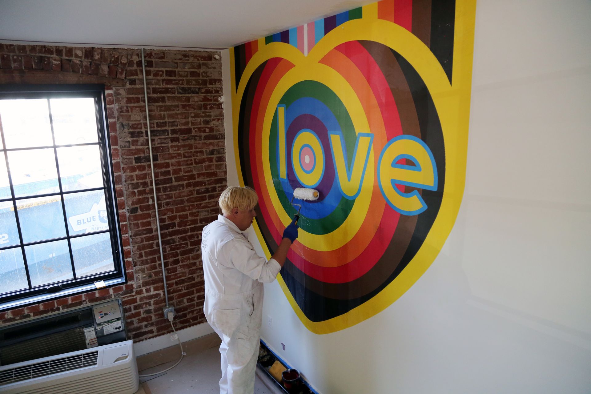 Person painting a colorful “love” mural on a white wall in a brick room