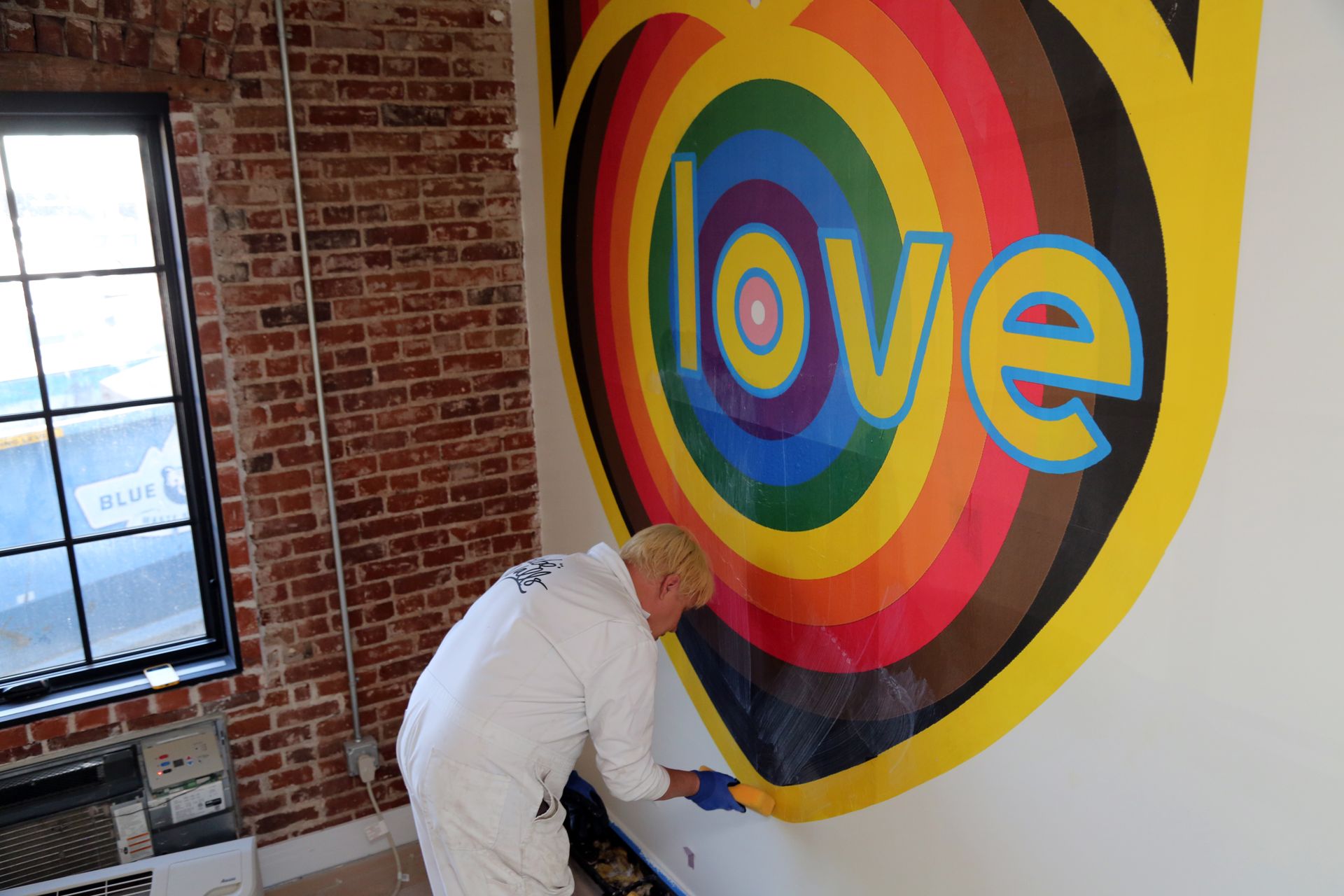 Person painting a colorful “love” mural on a white wall beside a brick window.
