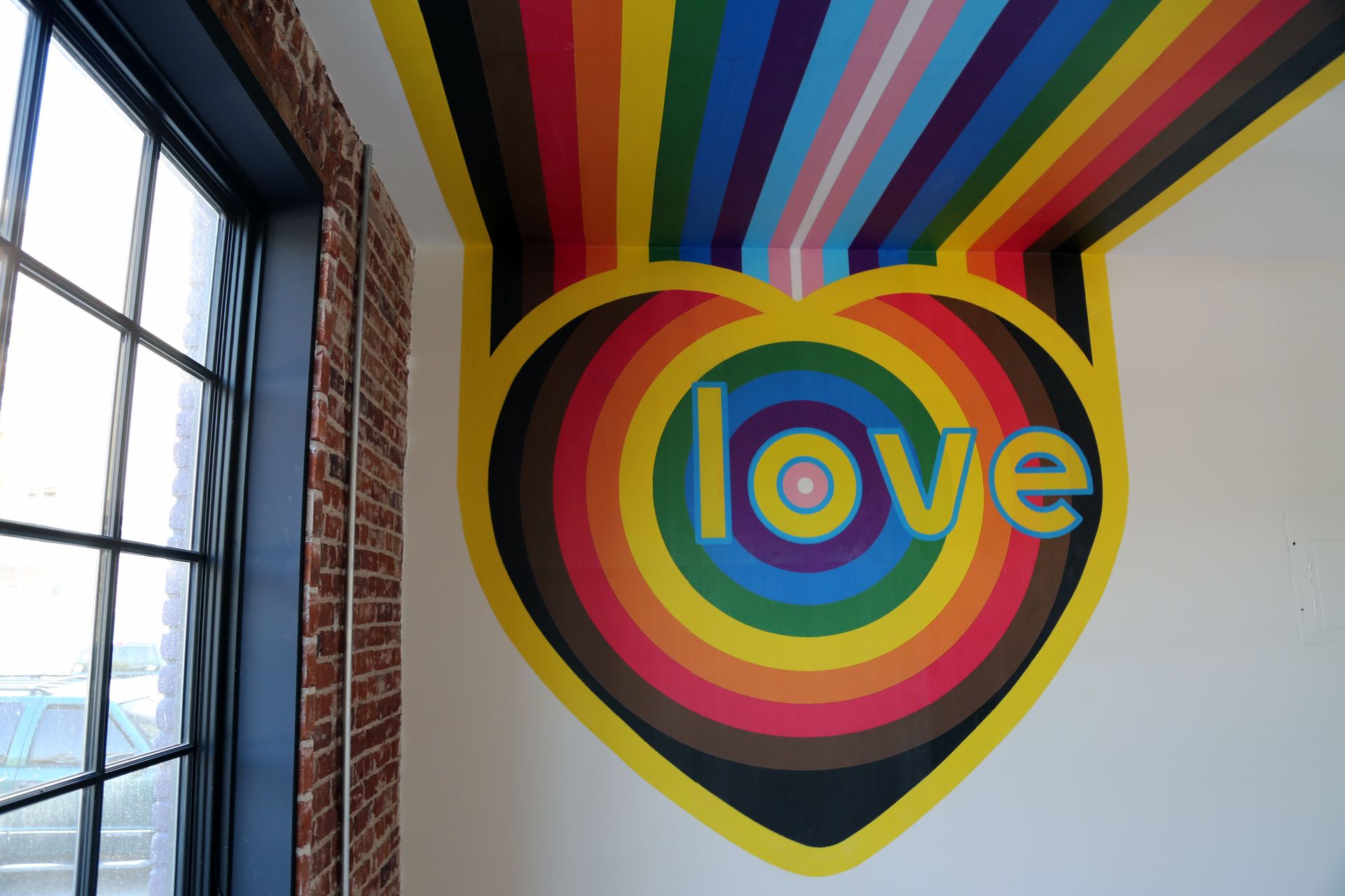 Colorful “love” mural with rainbow stripes on a white wall beside a window and brick column