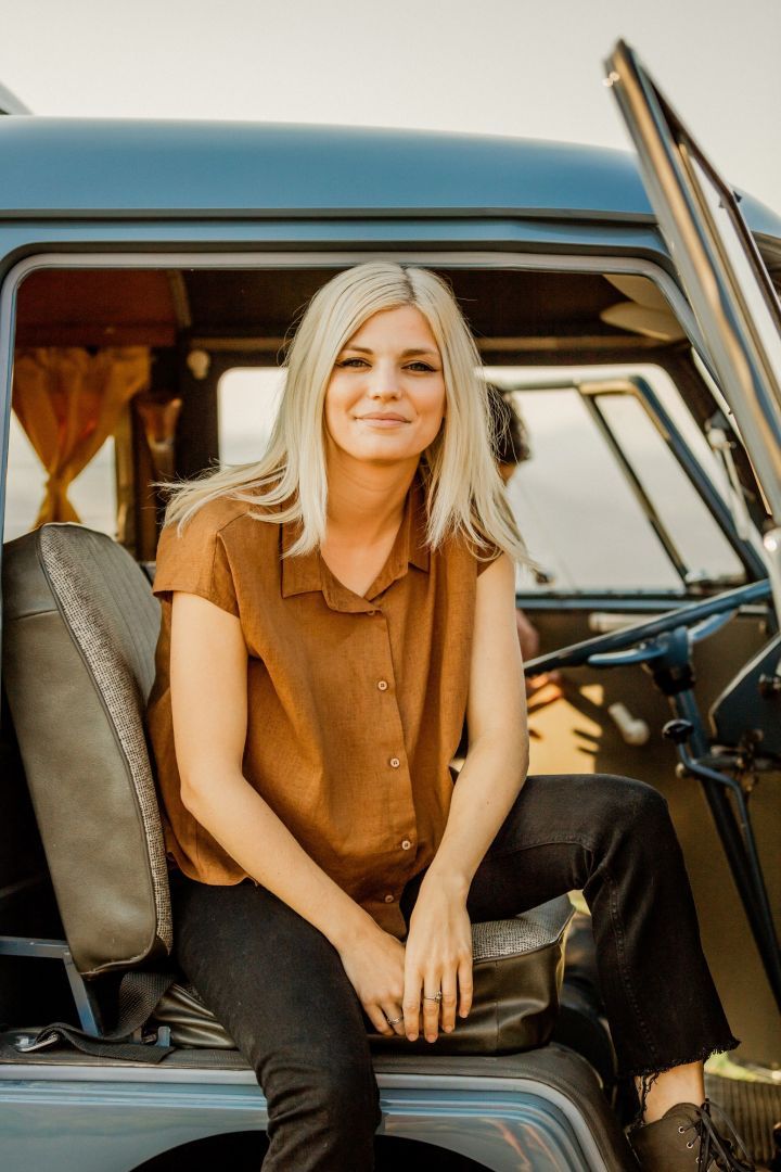 Blonde woman sitting in a vintage car doorway, wearing a brown sleeveless top and black pants.