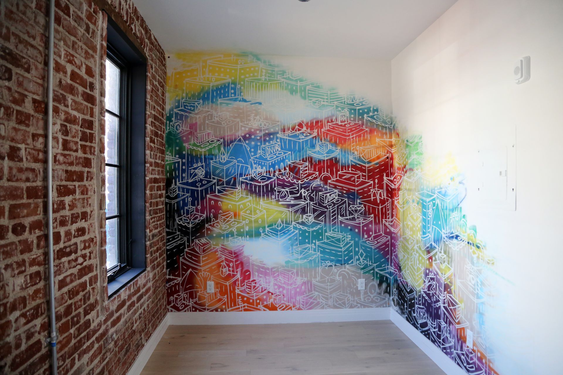 Empty room with a colorful abstract mural on the far wall, exposed brick on the left, and a window.