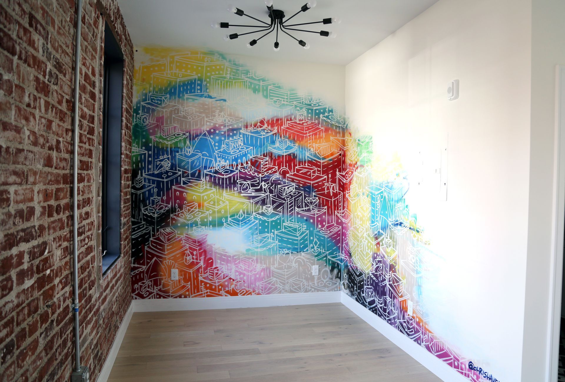 Small room with a colorful abstract mural on the back wall, exposed brick side wall, and wood floor.