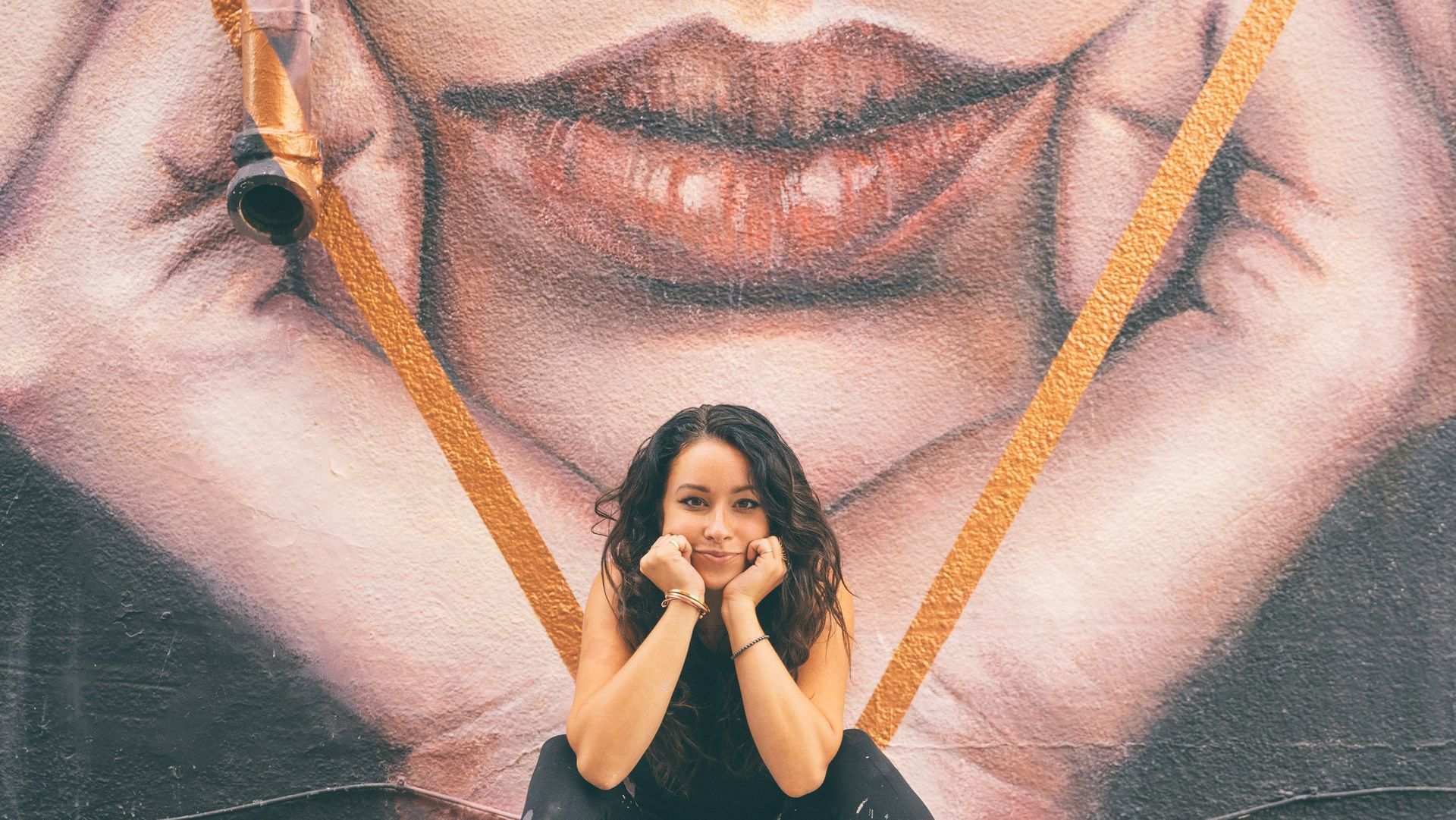 Woman posing in front of a mural with a large mouth and hands holding yellow strings