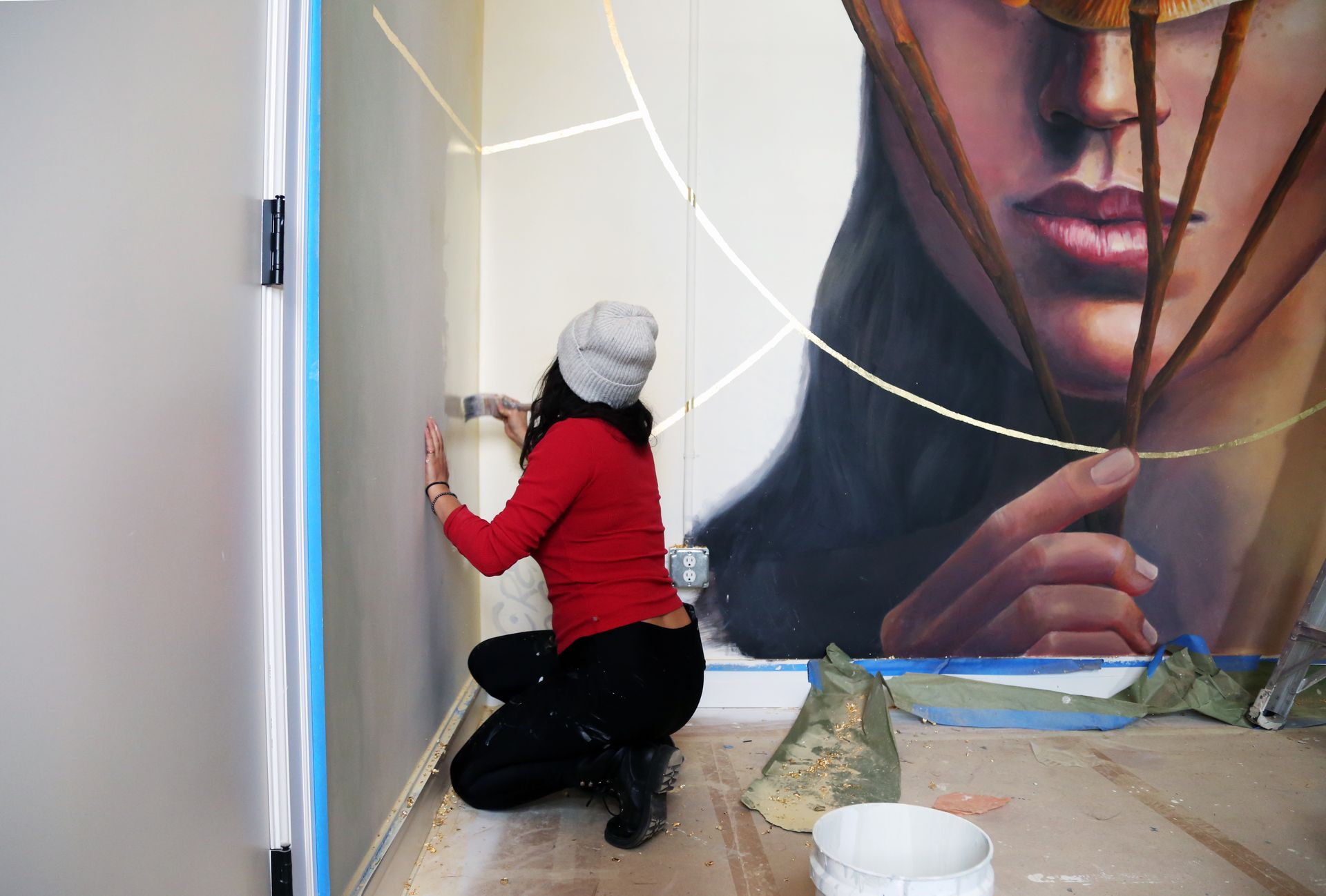Person in red hoodie painting a white wall beside a mural portrait with taped curves.
