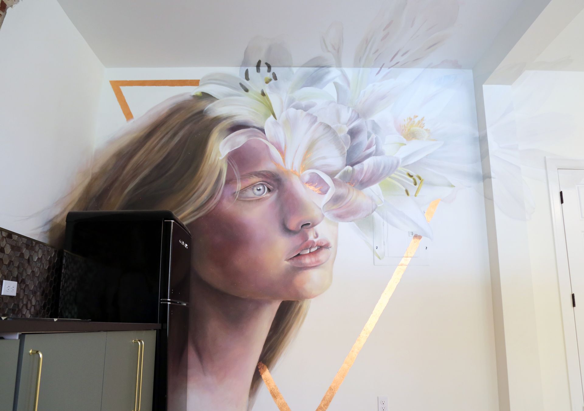 Large mural of a woman’s face with white flowers, painted on an indoor wall above a cabinet.