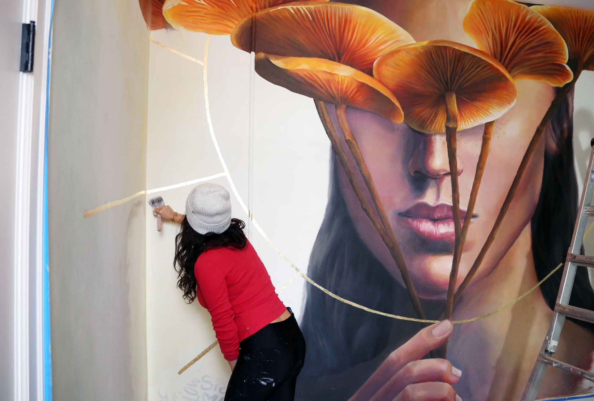 Person in red sweater reaching toward a mural of a face with large orange mushrooms above it
