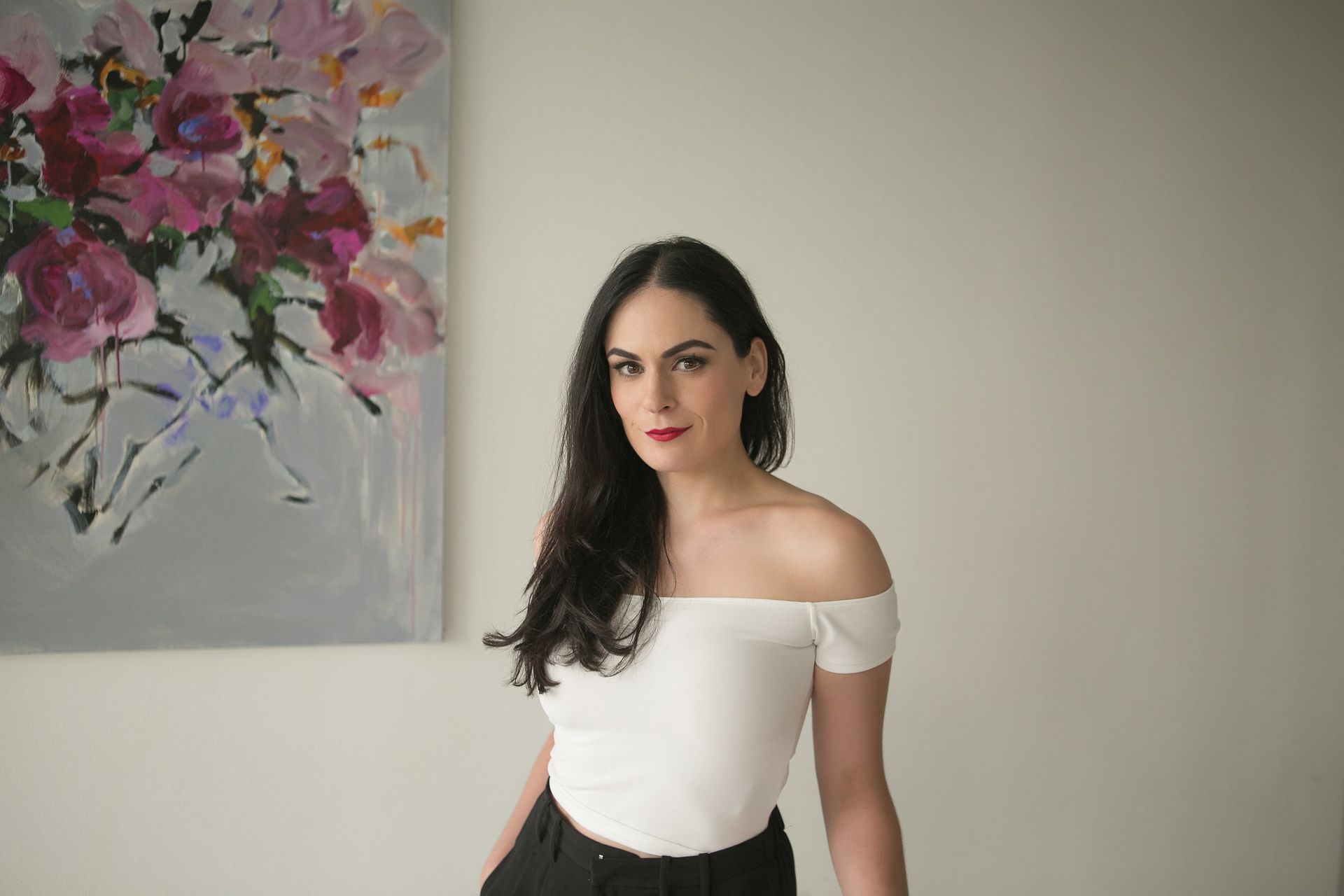 Woman in a white off-shoulder top standing beside a pink floral painting against a beige wall