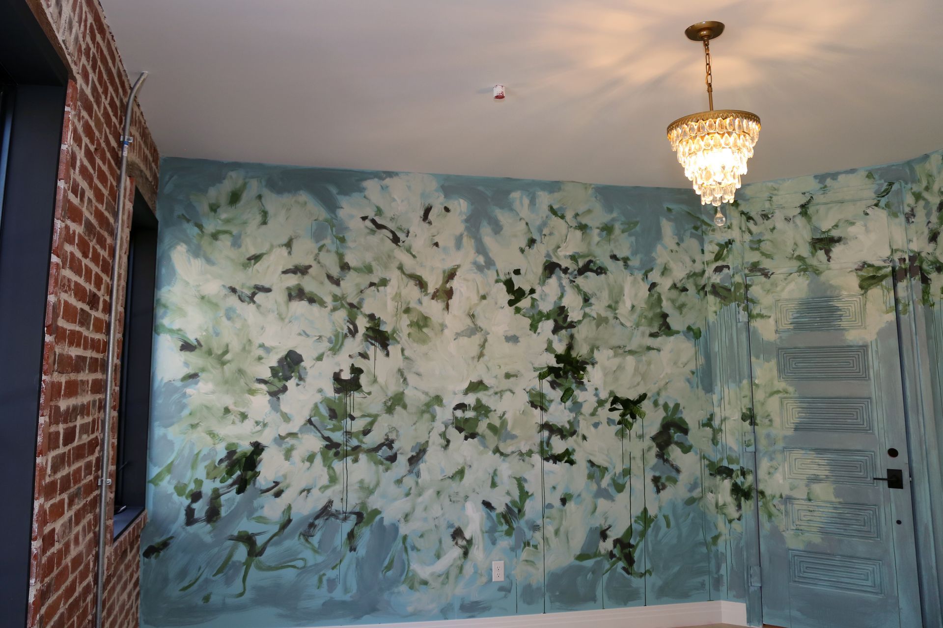 Floral mural wall in soft blue and white, with a chandelier and door in a hallway.