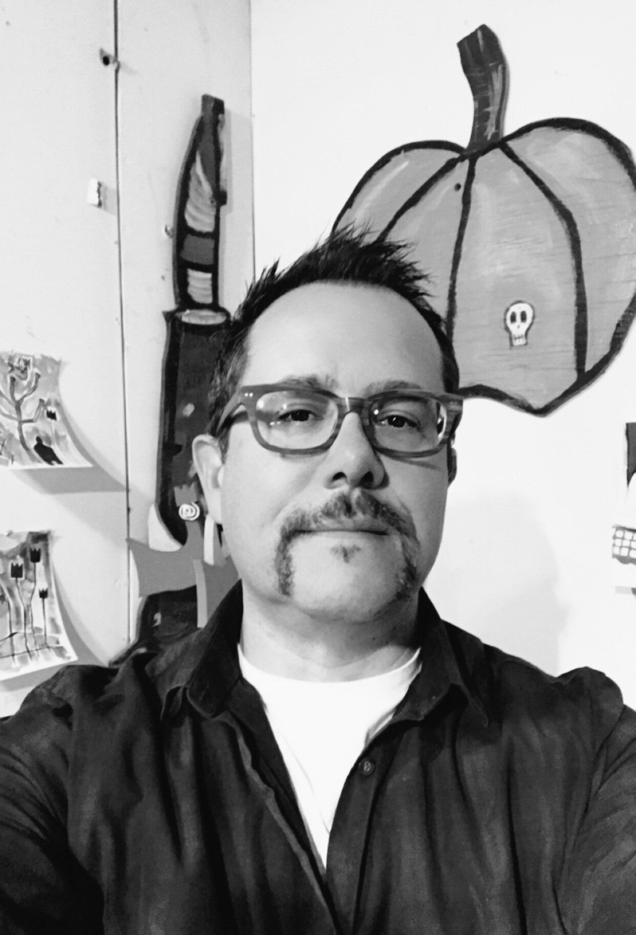 Man with glasses and mustache in a studio, wearing a dark jacket, with a pumpkin drawing in the background