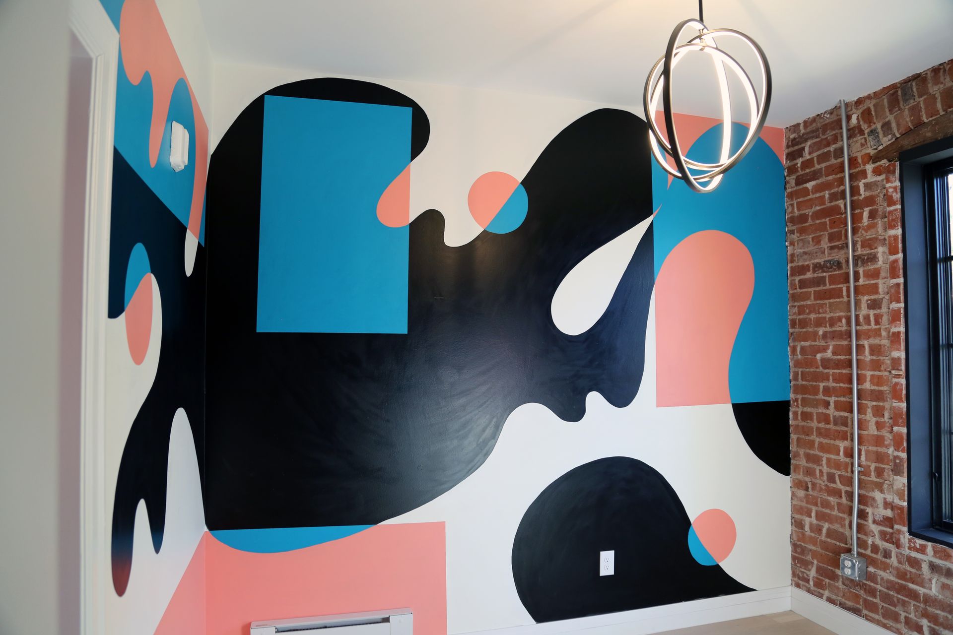 Abstract mural with black, blue, and coral shapes on a white wall beside exposed brick and a hanging light fixture