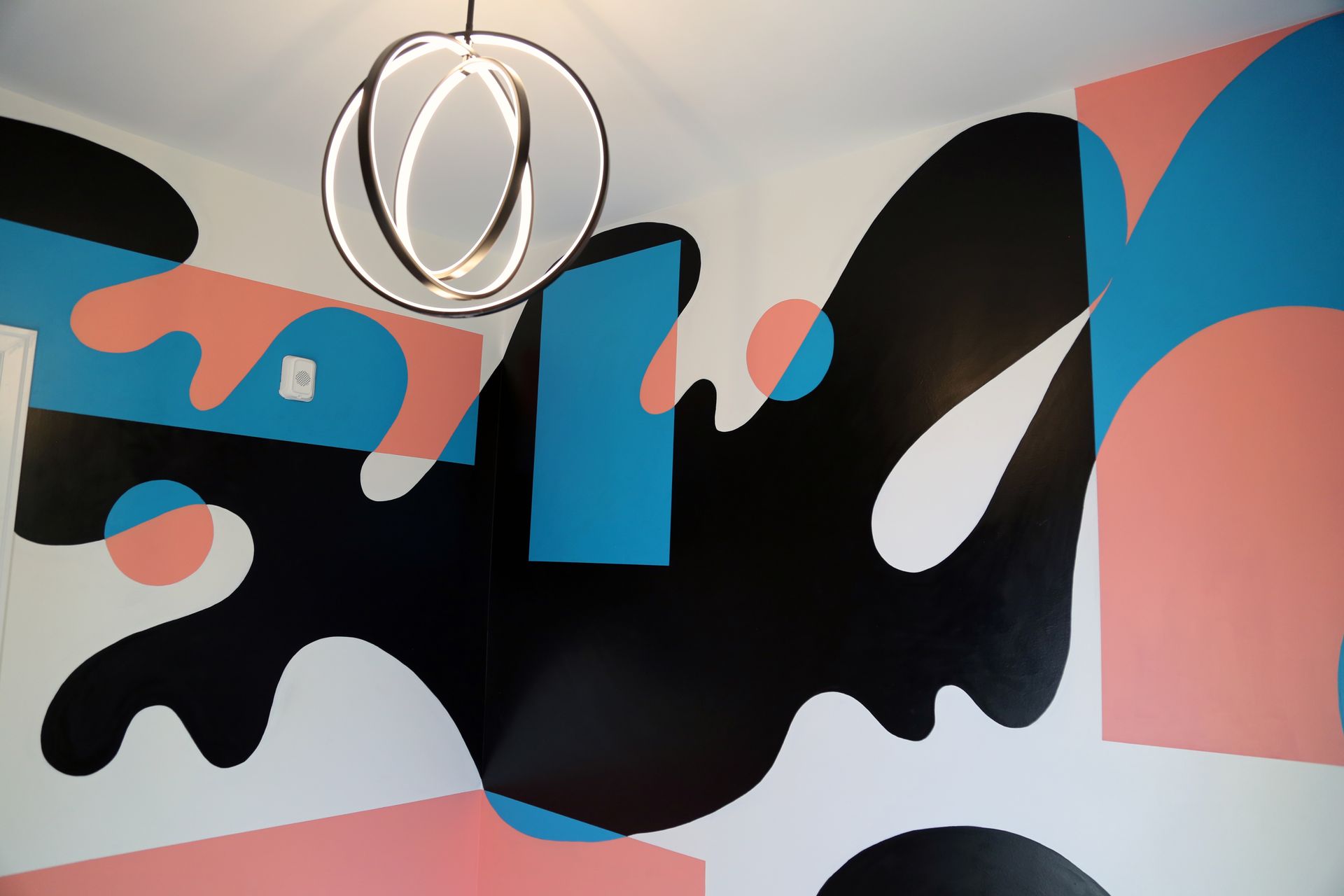 Abstract mural with black, blue, and pink shapes on a wall, with a hanging ring light above.