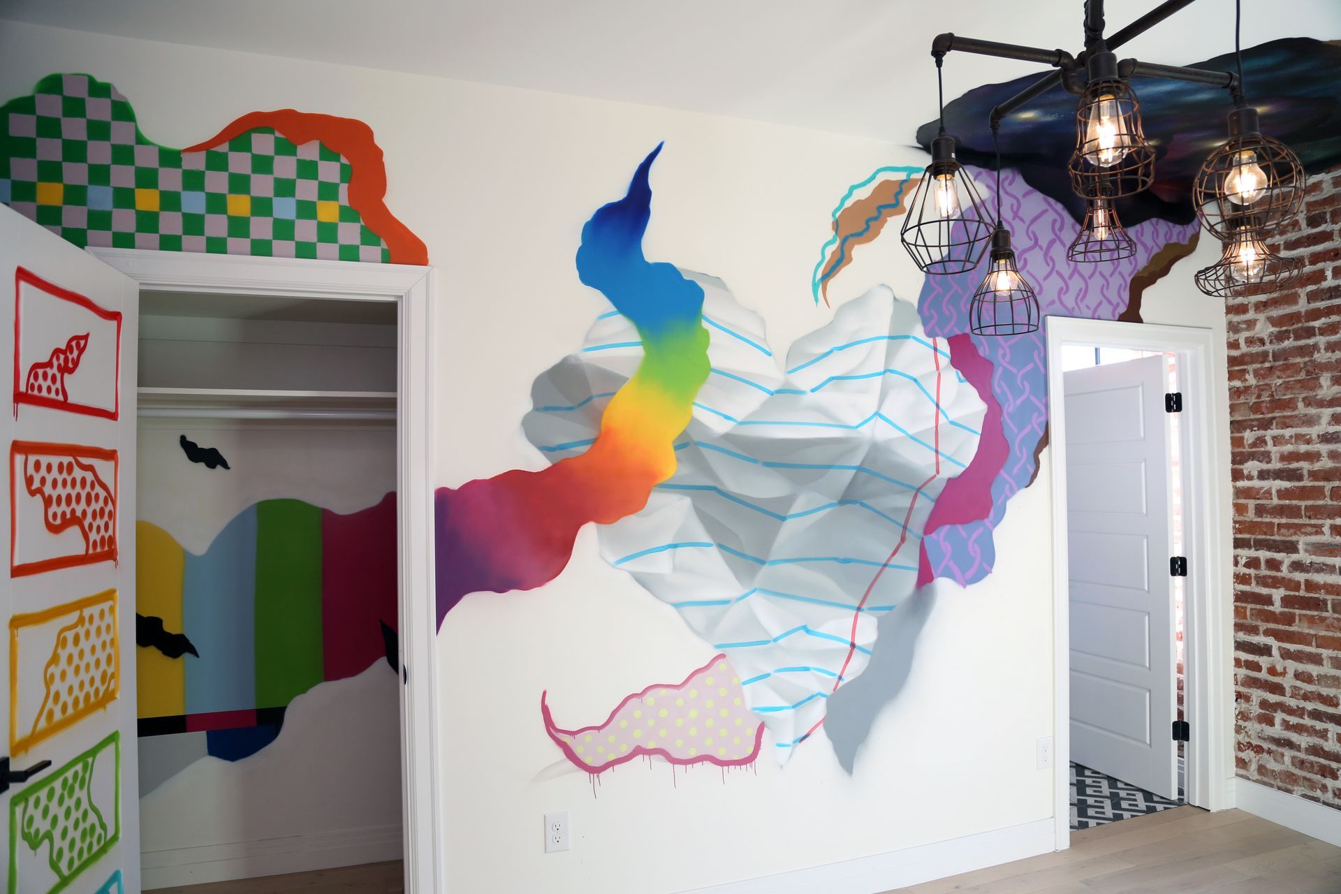 Colorful abstract mural on white gallery walls with black pendant lights and framed art near a doorway