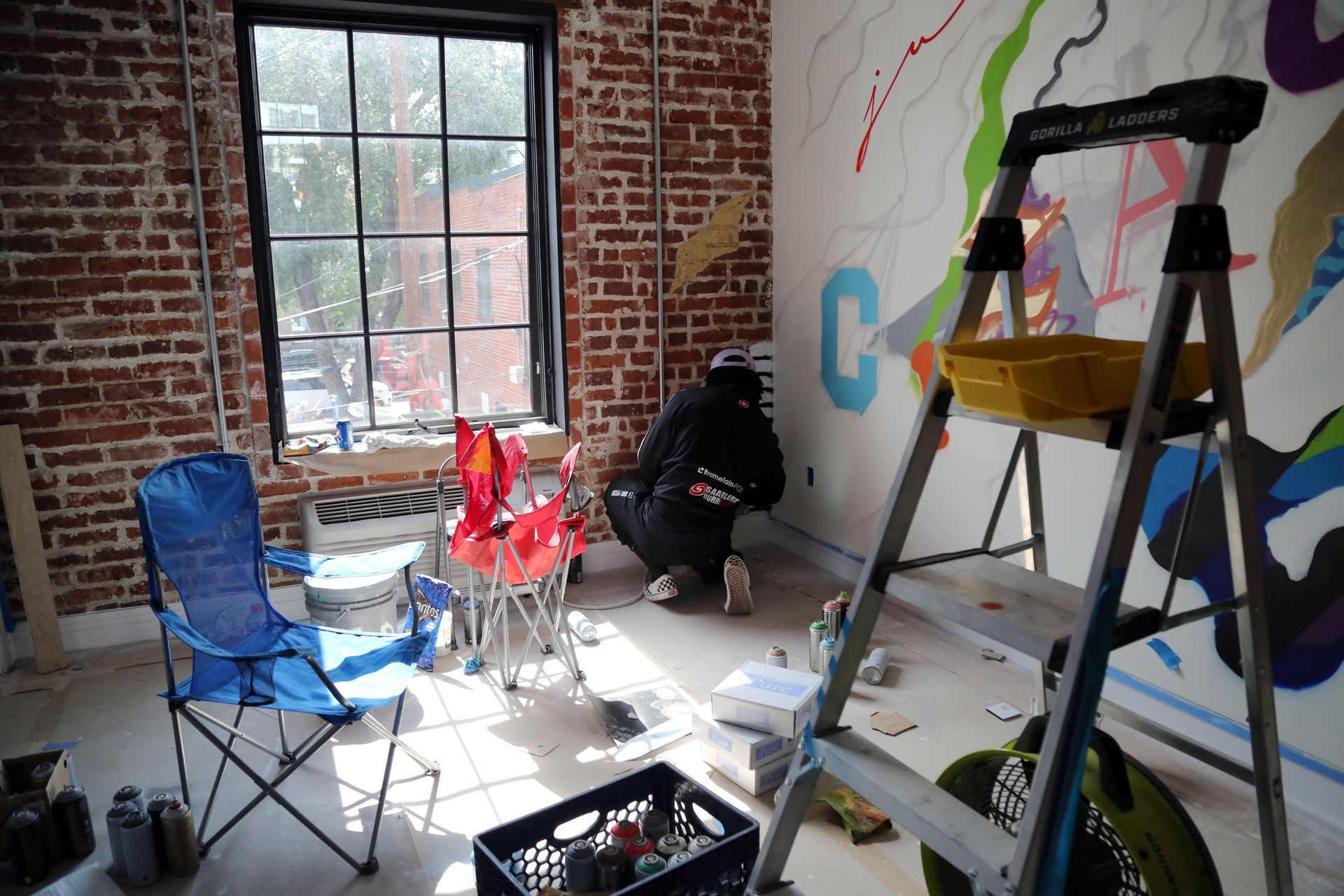 Sunlit room with exposed brick walls, folding chairs, a ladder, and a person crouching near the window