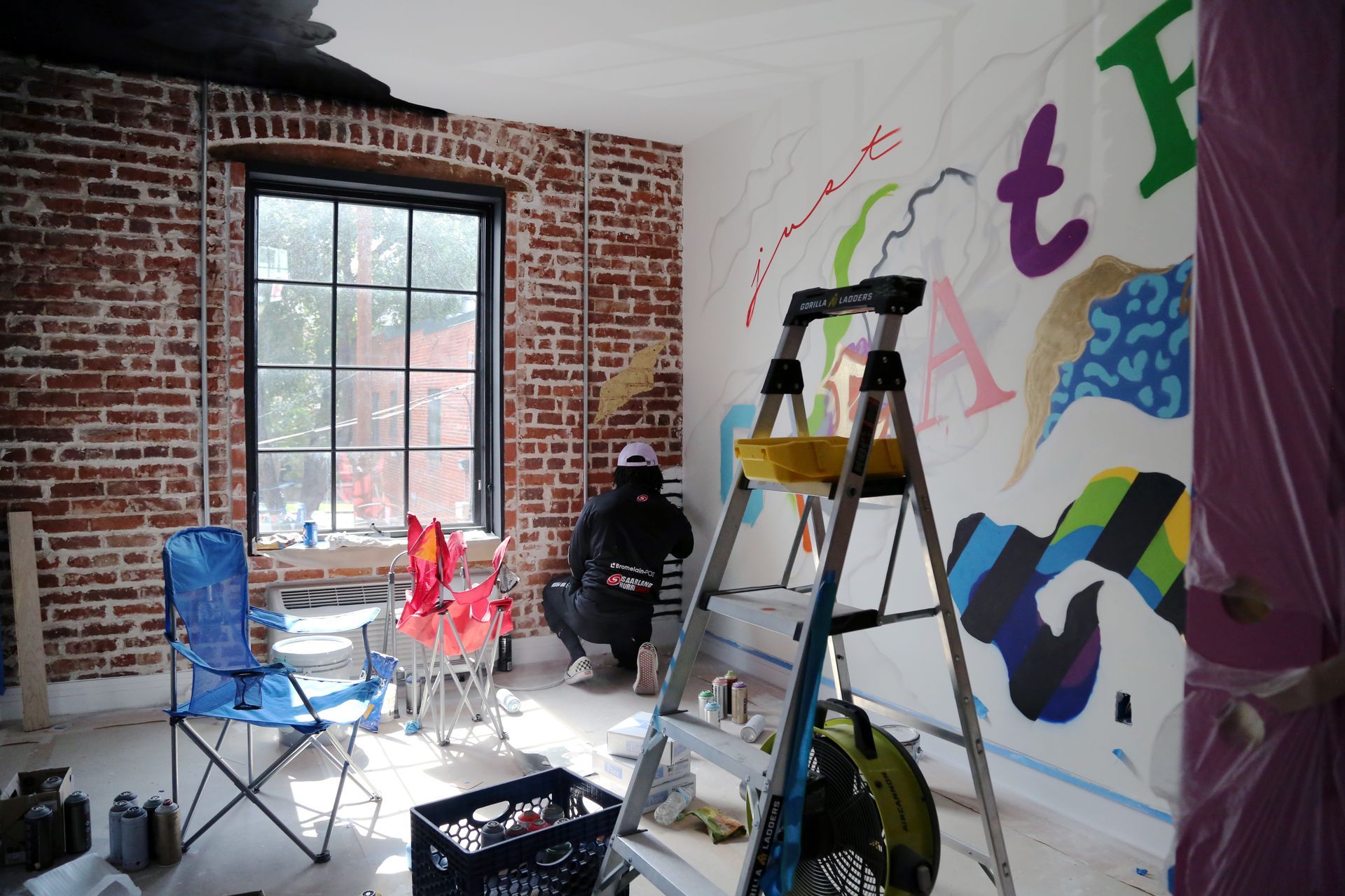 Person painting a brick-walled room with colorful mural, ladder, and tools scattered around.