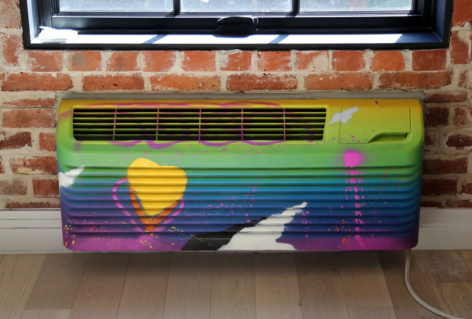 Colorfully painted radiator under a window against a brick wall, with a heart and abstract shapes