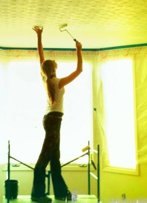 Person reaching up with a paint roller to paint a bright yellow ceiling in a room with tall windows.