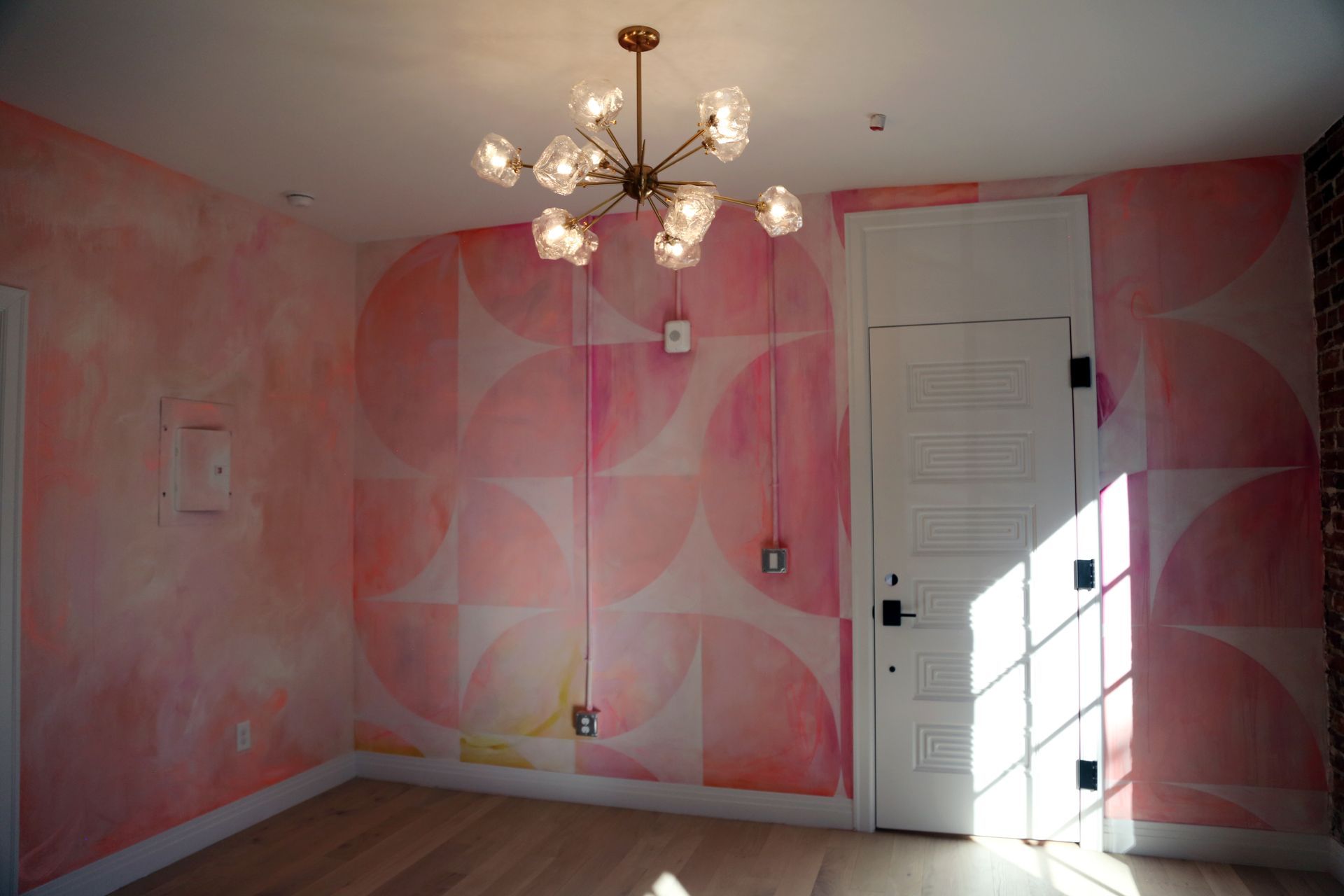 Empty pink-painted room with chandelier, white door, and sunlight on the floor
