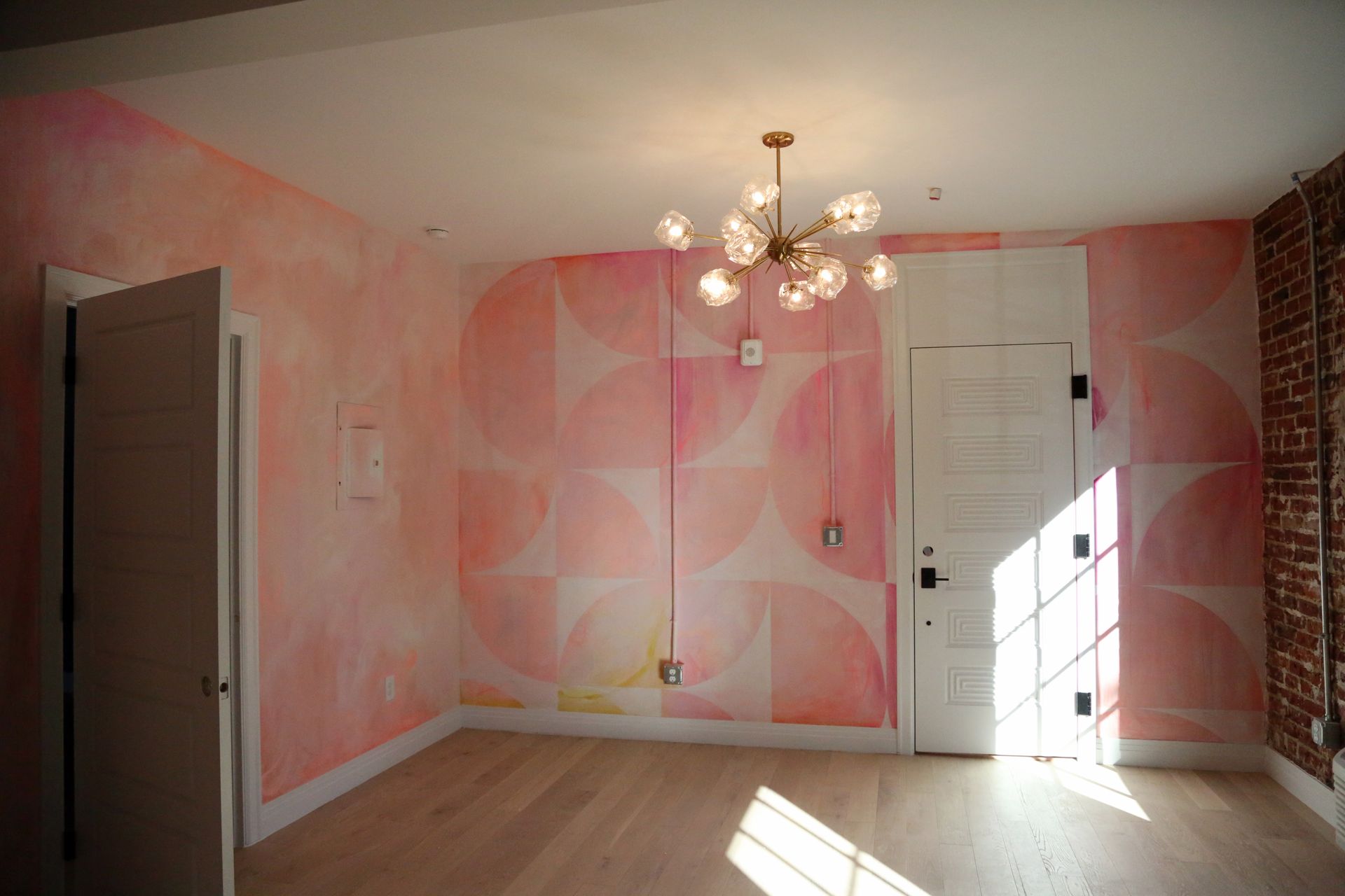 Empty pink-walled room with chandelier, white doors, and sunlight on hardwood floor