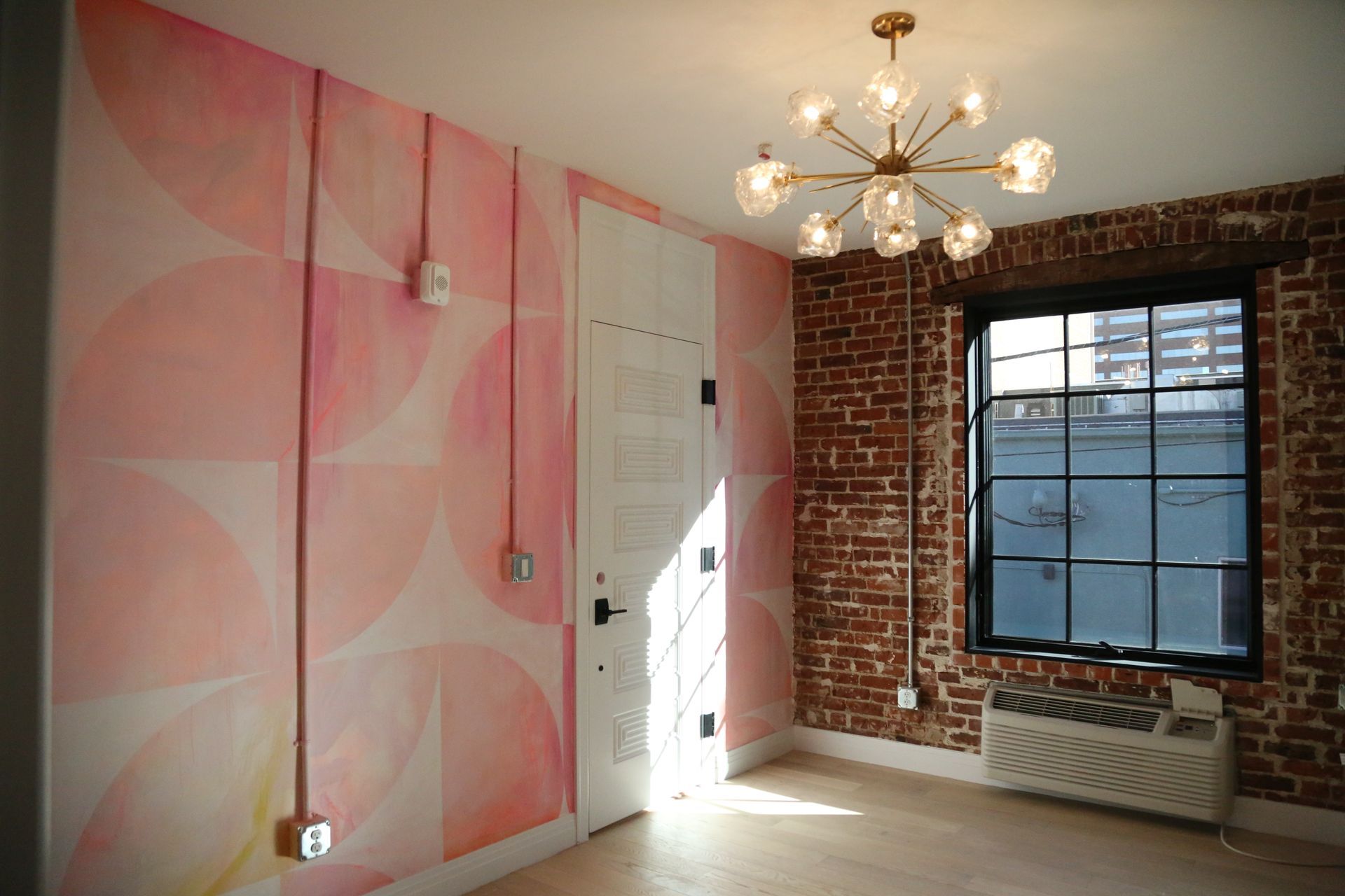 Small room with pink patterned wall, exposed brick, chandelier, and large window with radiator below.