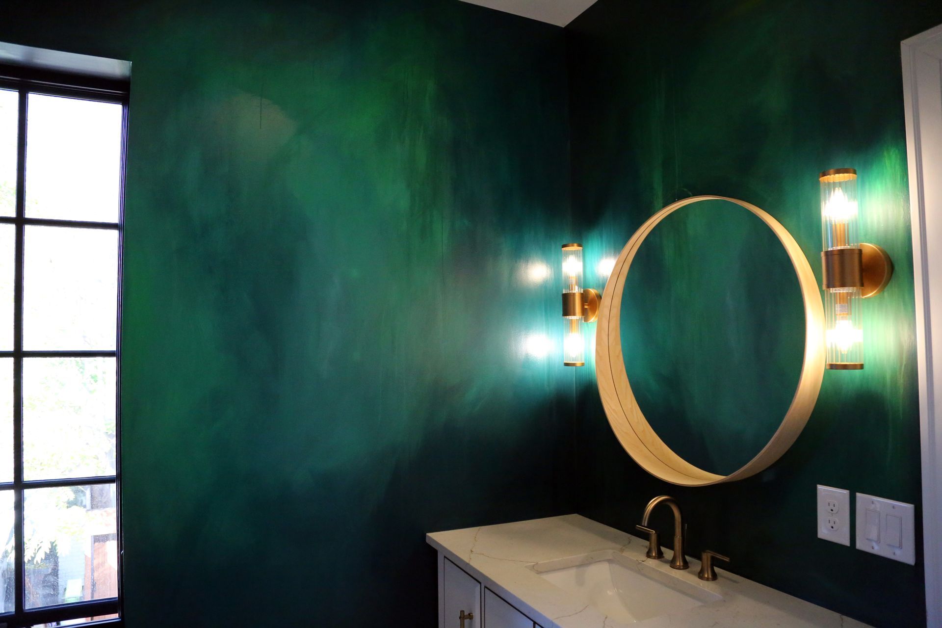 Bathroom vanity with round gold mirror, teal wall, lit sconces, and a window on the left.
