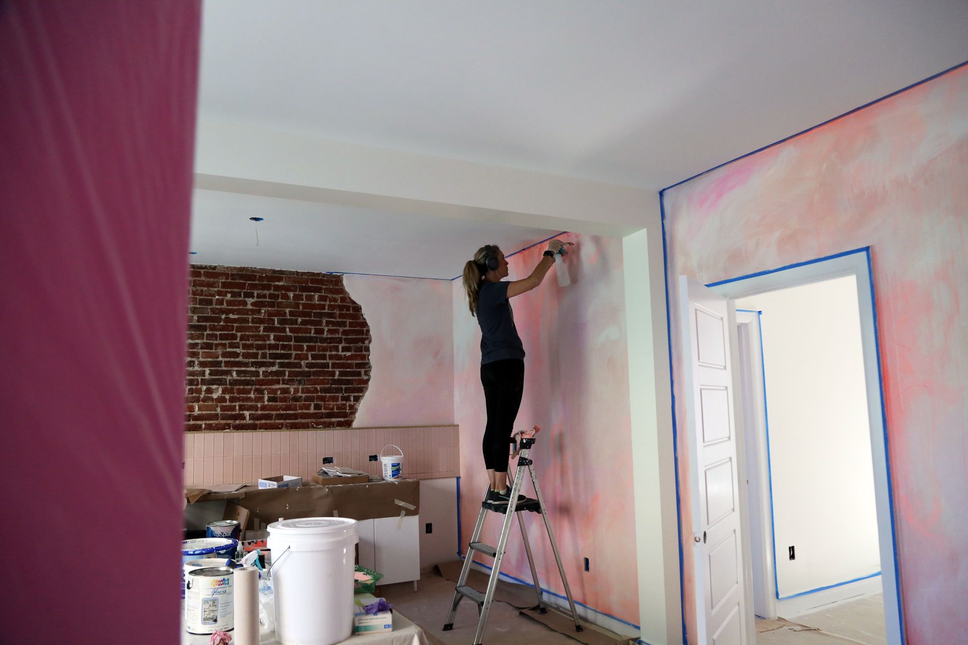 Person painting a pink wall indoors on a ladder in a room under renovation
