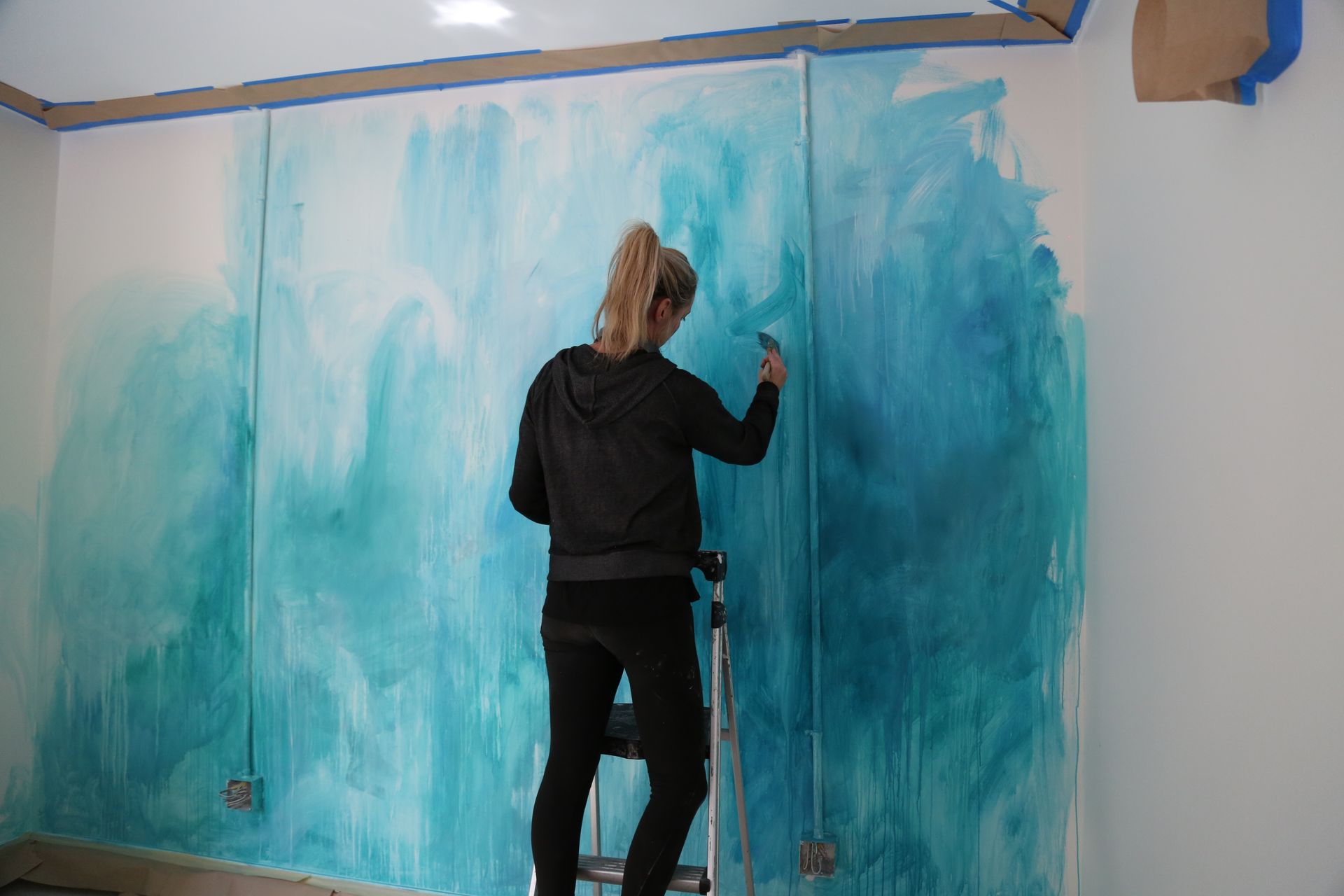 Person painting a wall teal and blue in a room, standing on a step stool