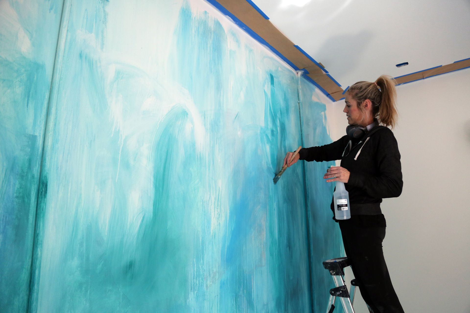 Woman painting a large blue mural on an indoor wall from a step ladder
