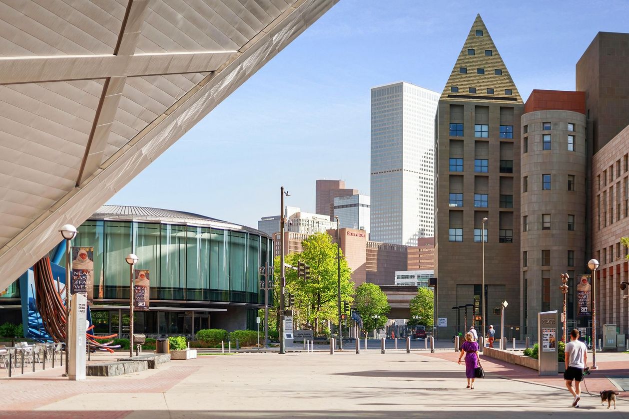 Modern plaza with pedestrians, a glass building, and a triangular-roofed tower under a bright sky