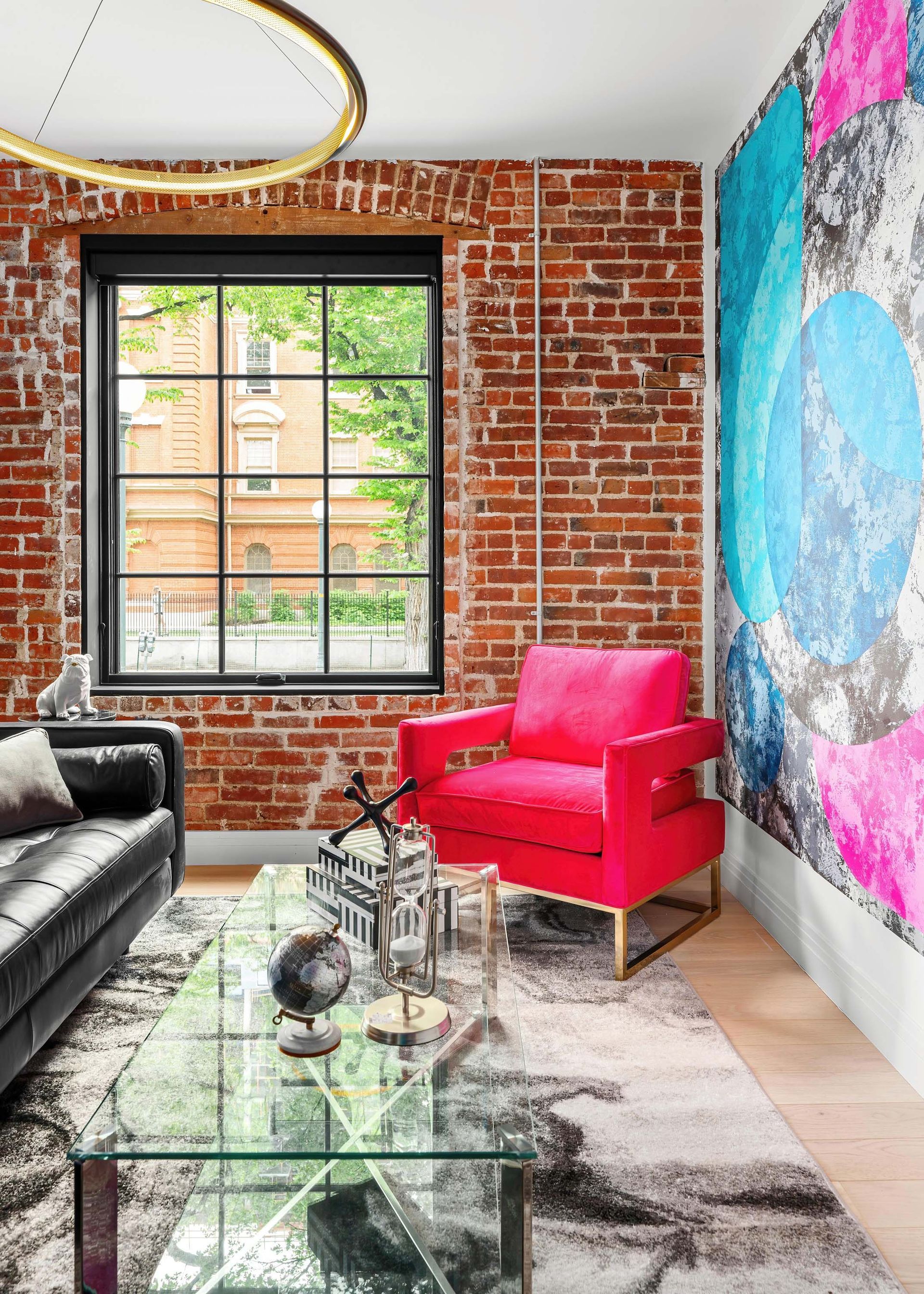 Bright loft living room with exposed brick, pink chair, black sofa, glass table, and colorful wall art.