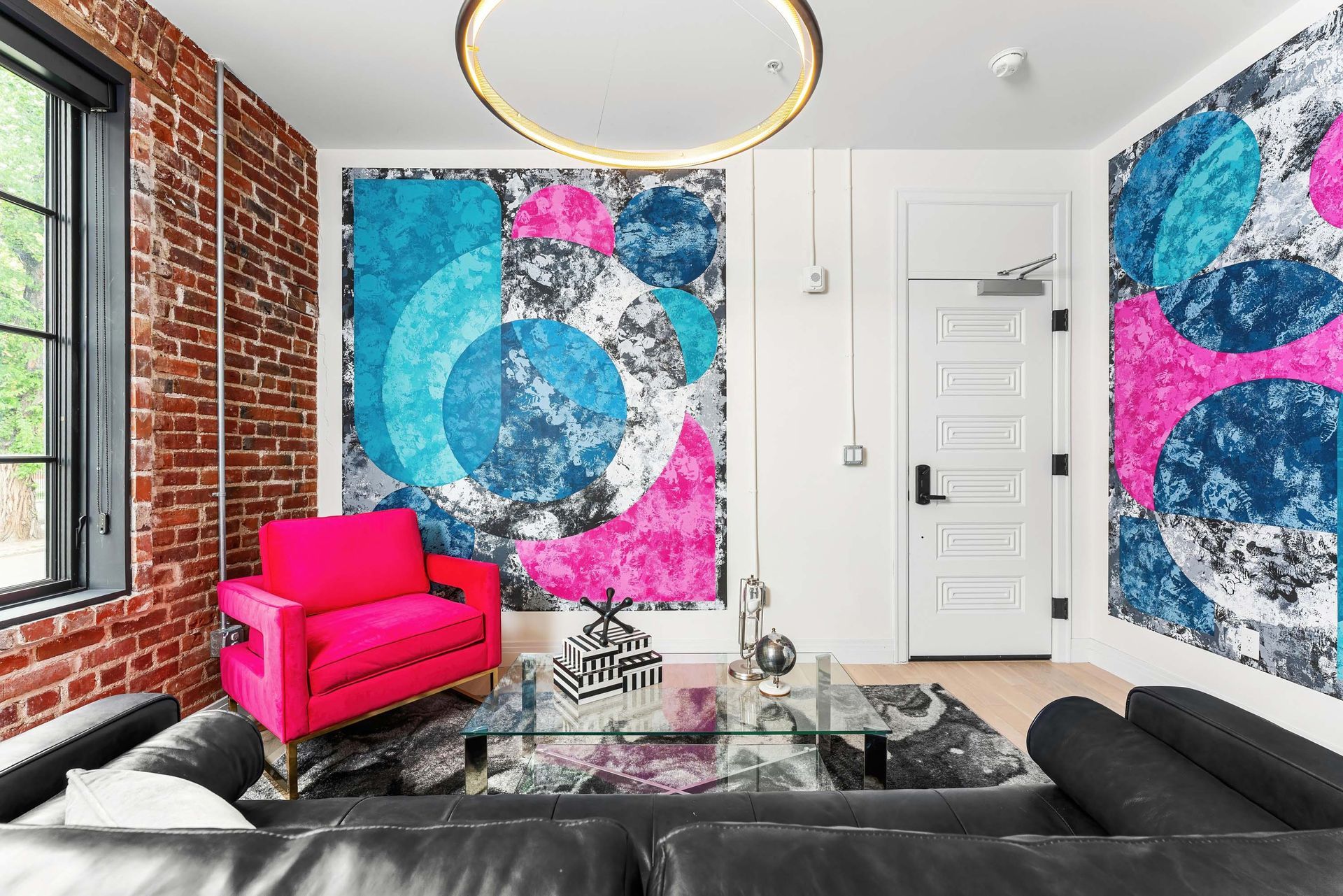 Modern living room with bold abstract wall art, pink chair, black sofa, and exposed brick wall