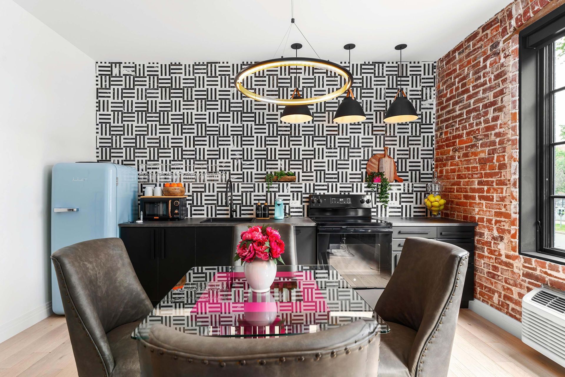 Modern dining kitchen with patterned wallpaper, brick wall, black cabinetry, and a round table with pink flowers.