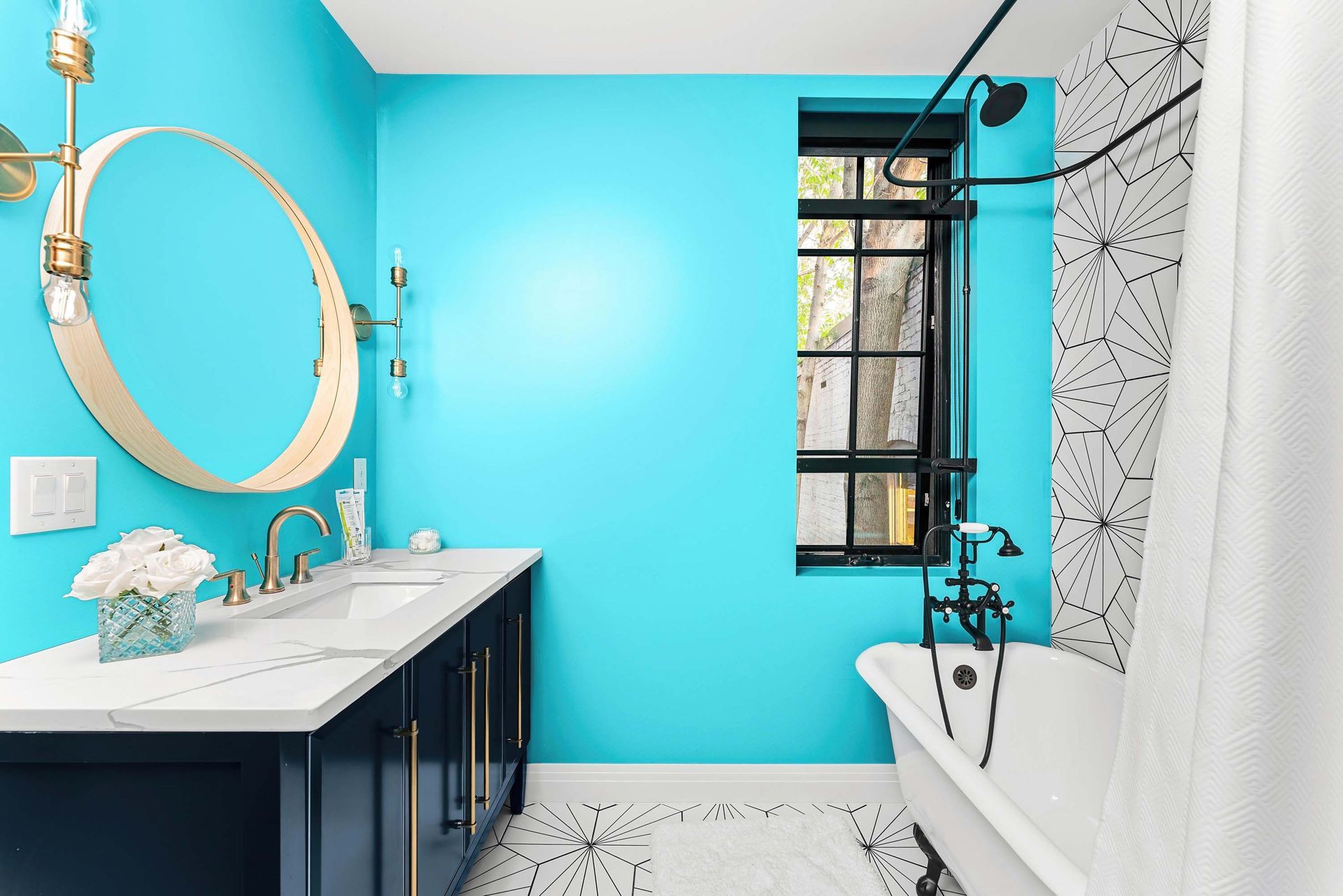 Bright turquoise bathroom with white vanity, round mirror, and black-framed window beside a bathtub.